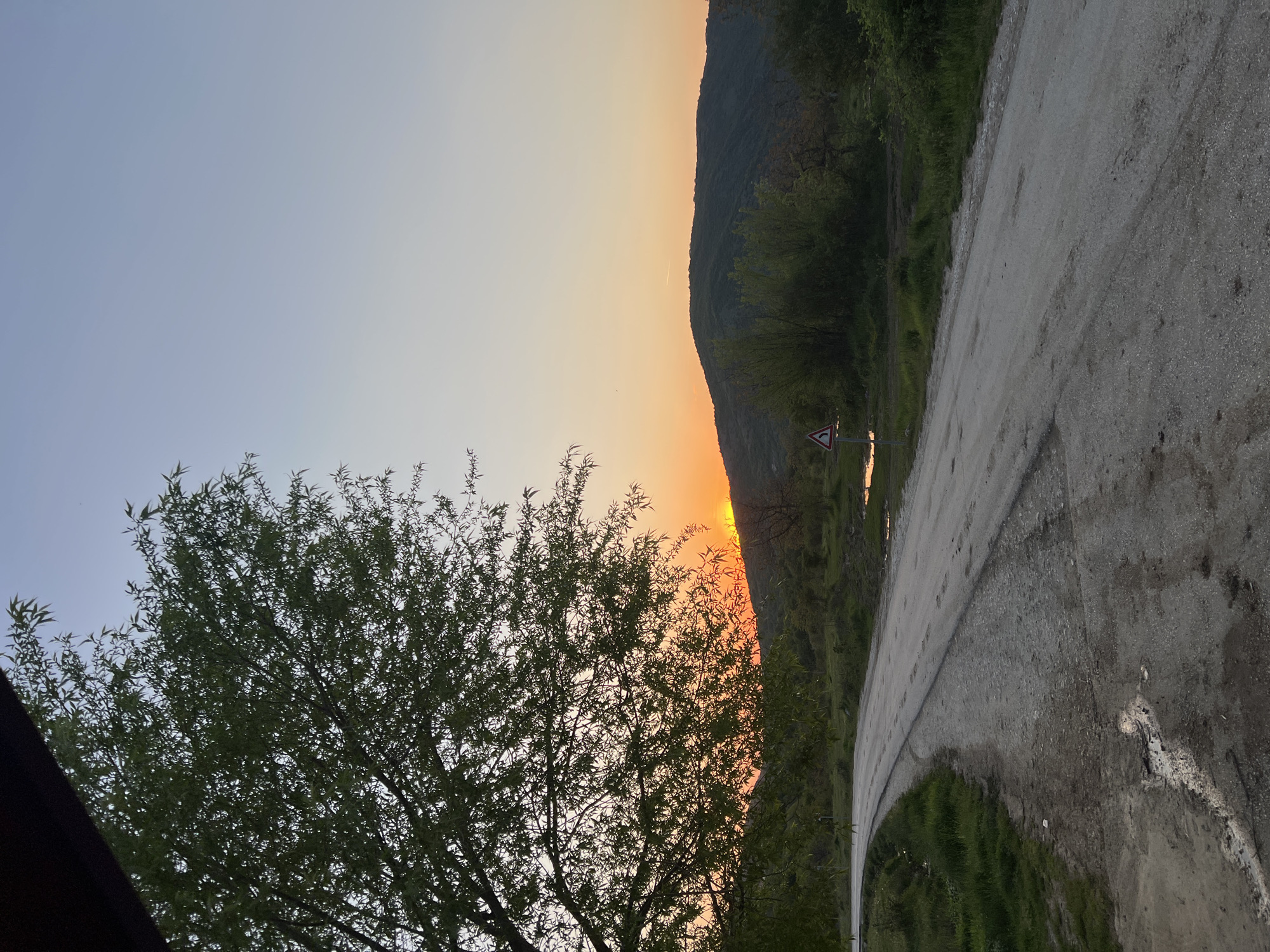 The image shows a rural road in Galabovo, Bulgaria, at sunset. The road curves to the right, with a hillside on the right side of the frame. A road sign indicating a curve is visible. A tree is on the left side of the frame, partially obscuring the sunset. The sky transitions from blue to orange as the sun sets behind the hill.