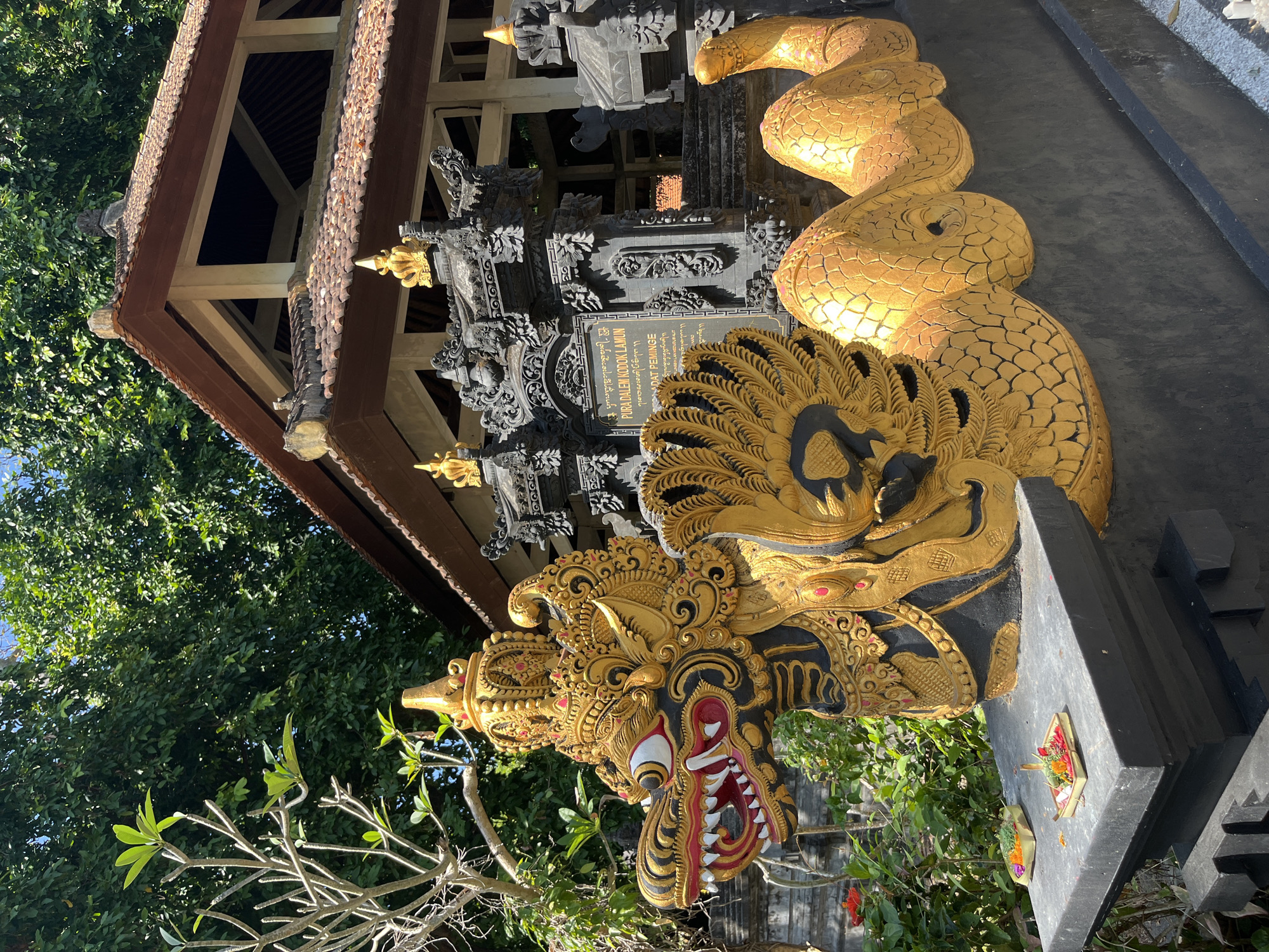 The image shows a detailed, golden dragon sculpture with black accents, positioned in front of a traditional Balinese temple. The sculpture is ornate, featuring a crowned head with prominent red and white eyes, sharp teeth, and a long, scaled body that coils to the right. On a black stone pedestal in front of the sculpture, two small offerings are placed, consisting of flowers and incense. Behind the dragon, a stone temple gate with intricate carvings is visible, leading to a multi-tiered building with a reddish-brown tiled roof. Lush green trees form the background, suggesting an outdoor, tropical environment. The location is identified as Pura Dalem Kodok Lamun, which is situated in the city of Kuta Selatan, Indonesia. The scene is brightly lit by sunlight, casting shadows and highlighting the vibrant colors and textures of the sculpture and temple.