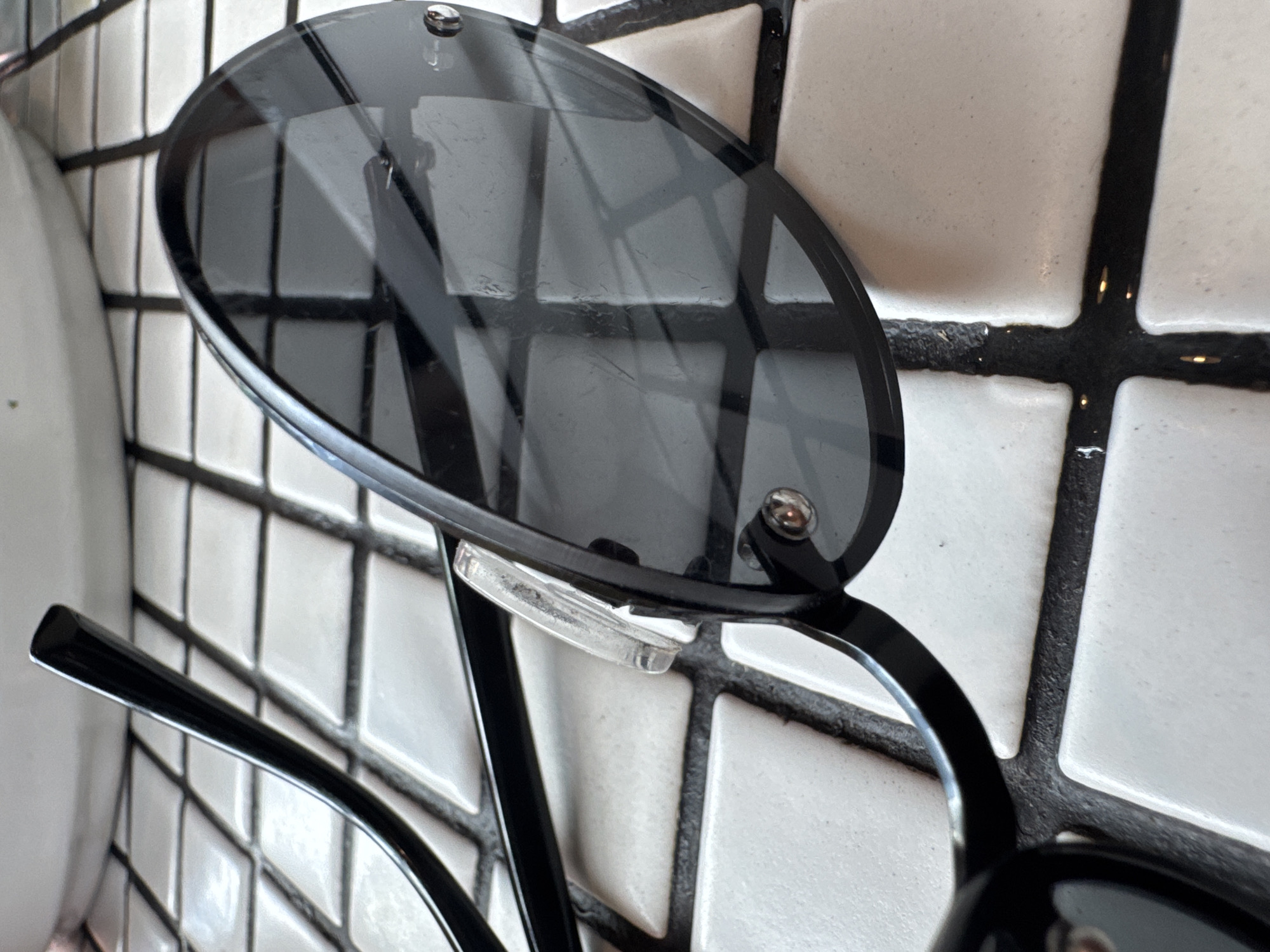 A close-up shot shows a pair of black-rimmed sunglasses resting on a tiled surface, likely in an indoor setting. The tiles are white with black grout, forming a grid pattern. The left lens of the sunglasses is positioned prominently, revealing its dark tint and a few minor scratches. A clear nose pad is visible between the lenses. The sunglasses are in focus, while the background tiles are slightly blurred. The image suggests a casual, everyday scene. The location, North Kuta, Indonesia, is not directly indicated within the image itself.