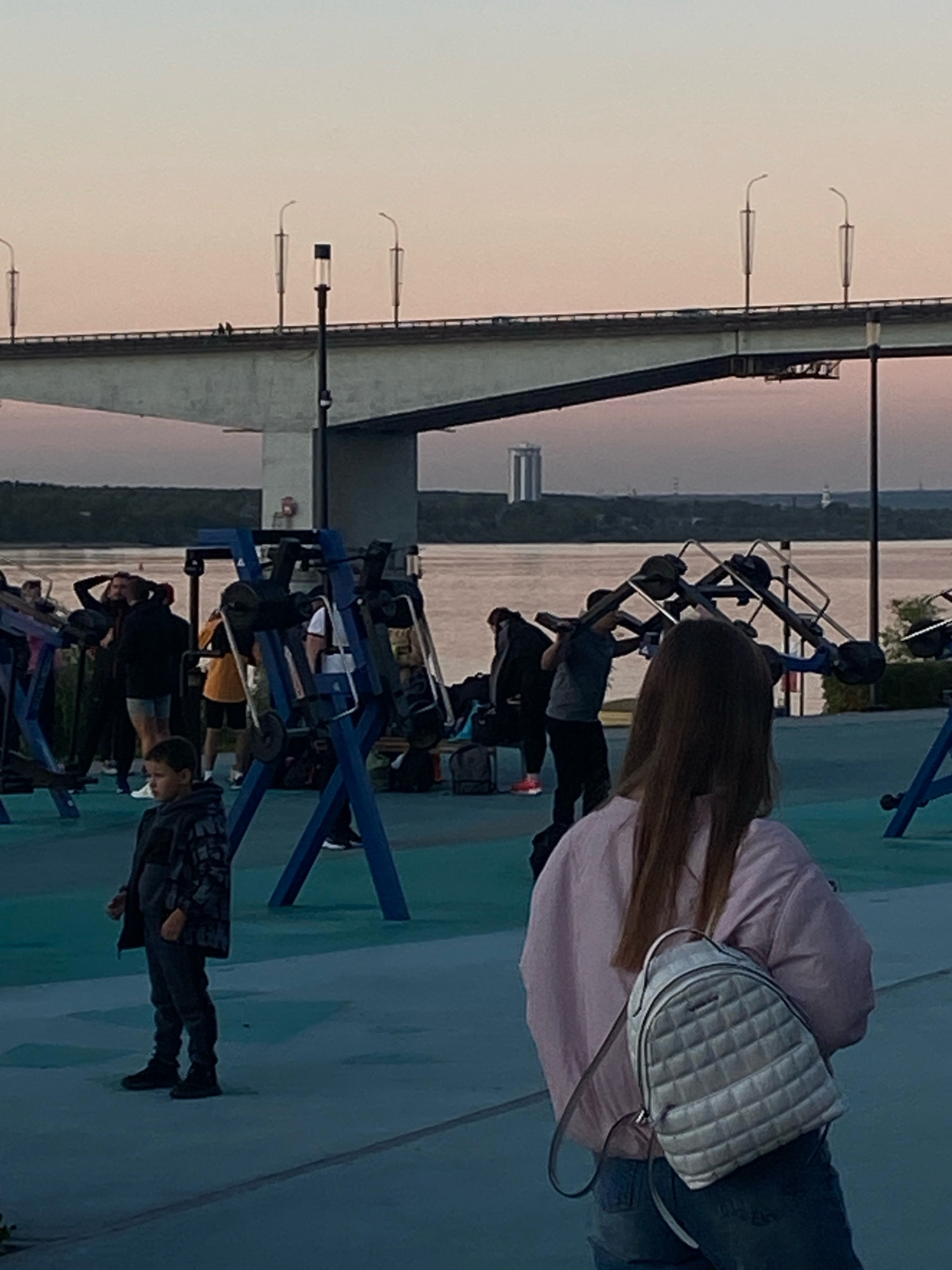 The image depicts an outdoor scene near the Kama River embankment in Perm, Russia, with a bridge spanning the water in the background. In the foreground, a young boy in a patterned jacket and dark pants stands on a blue-toned paved surface. To his right and further back, a woman with long brown hair wears a light pink jacket and jeans, carrying a quilted silver backpack. Several other individuals are visible in the middle ground, some appearing to be engaged in exercises on outdoor gym equipment. The overall atmosphere suggests a late afternoon or early evening setting, with soft lighting and a muted sky. The environment includes elements of urban infrastructure, recreational facilities, and natural landscape.