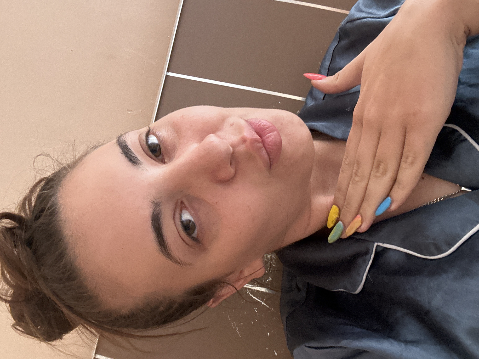 A woman with brown hair pulled back in a messy bun is posing for a selfie-style photo. She is wearing a silky, dark-colored top with white piping. Her fingernails are painted in various pastel colors. She has light skin, brown eyes, and full lips. The background appears to be a wall with a two-tone color scheme, possibly beige and brown with a silver accent strip. The photo does not contain information to link it to the city of Sirakovo, Bulgaria.