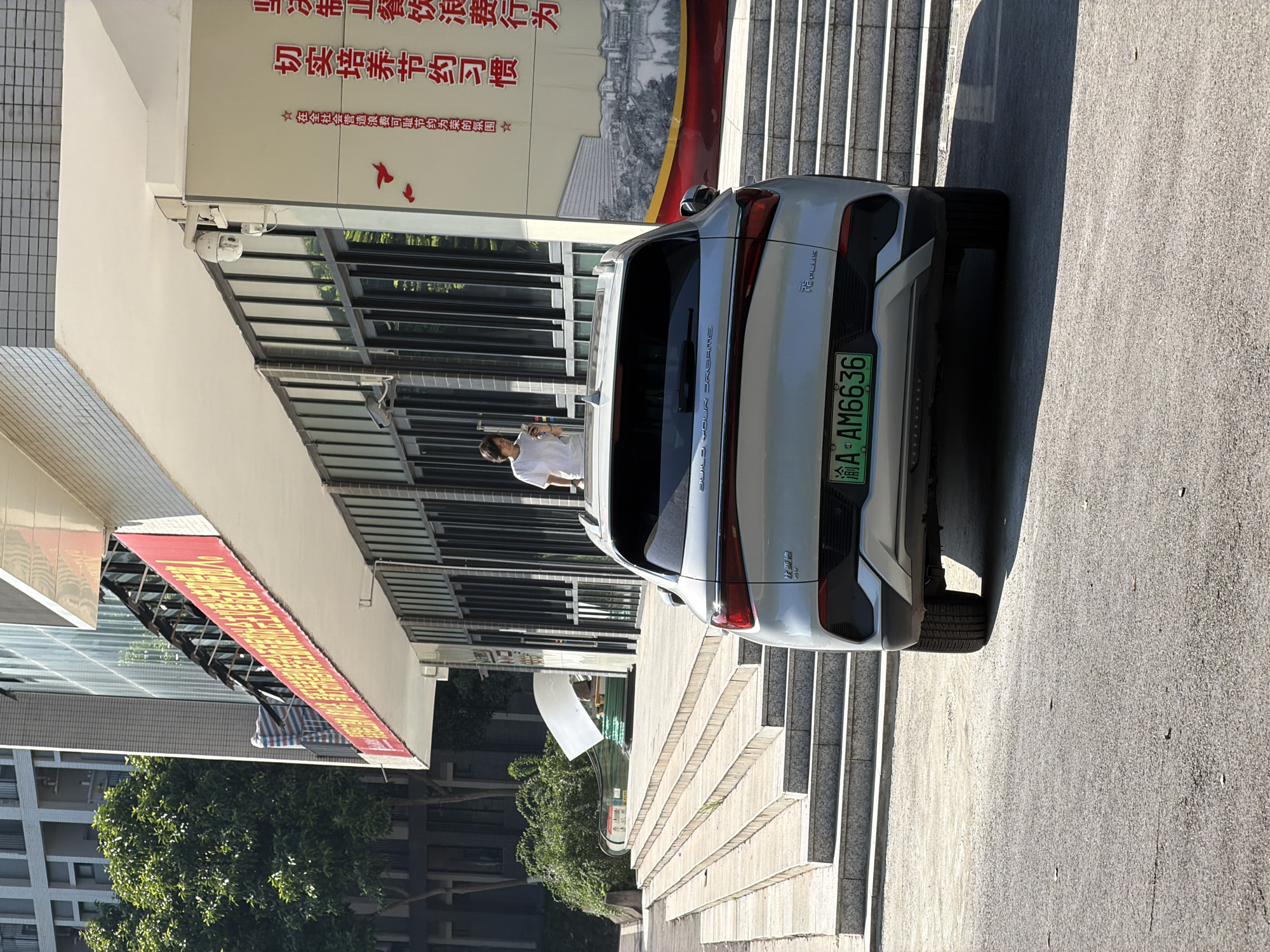 The image depicts an outdoor scene in the city of Jiangjin, China, on a sunny day. In the foreground, a silver SUV is parked on an asphalt surface. The rear of the vehicle is visible, displaying a green license plate with the characters "A-AM6636". The SUV has "FE PLUS" badging on its rear. To the left of the vehicle, a wide set of concrete stairs leads up to a building. A person wearing a white t-shirt is standing on the upper landing of these stairs, looking at their phone.
The building facade features a light-colored overhang, and large glass windows run along the ground floor. A red banner with Chinese characters is draped across the upper part of the building. Red decorative birds are also visible on the building's surface. To the left of the building, lush green trees are present, and a multi-story residential building is partially visible in the background. A security camera is mounted on the building's exterior. The overall environment suggests a campus or public facility.