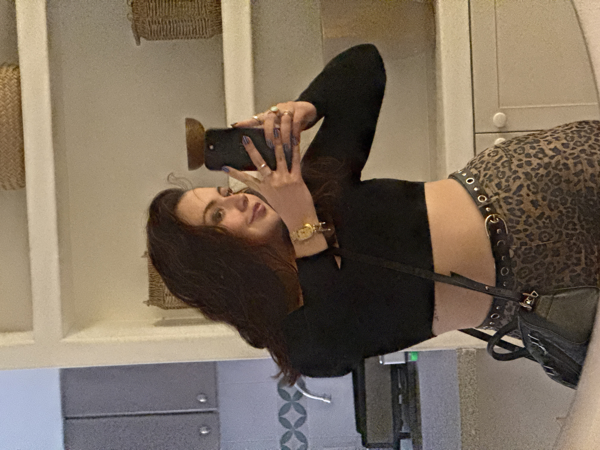 A woman is taking a selfie in a bathroom mirror. She is wearing a black long-sleeved crop top and leopard-print pants with eyelet detailing. A black purse hangs over her shoulder. She has dark hair, a watch on her left wrist, and multiple rings on her fingers. The bathroom has white cabinets and drawers, with decorative baskets on shelves. A wall with blue and white tile detail is visible behind her. The location is likely in Mexico City, Mexico, but there is no location-specific information visible in the image to confirm this detail.