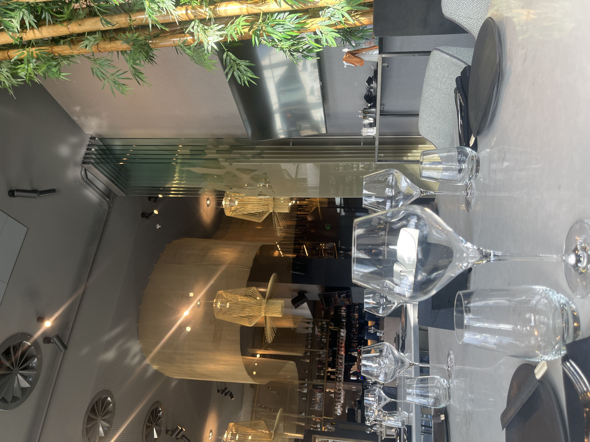 The image depicts the interior of a restaurant in Sofia, Bulgaria. The shot is taken from a table set with multiple wine glasses, plates, and silverware, suggesting a formal dining setting. The decor includes bamboo-like foliage overhead and modern lighting fixtures. The reflection in the glasses reveals a glimpse of the restaurant's interior, showing a bar area stocked with bottles and other seating arrangements. The color scheme is predominantly neutral, with grays and whites dominating the scene. No people are directly visible in the frame.