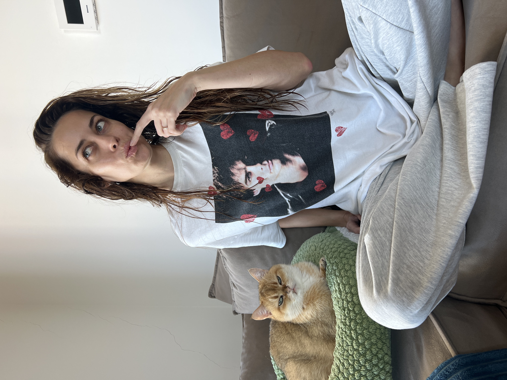 This image depicts an indoor residential setting in Bar, Montenegro, featuring one adult female and one domestic cat.
The female is seated on a multi-cushioned, neutral-toned sofa, with her legs crossed. She has medium-length, wet brown hair and a light complexion. She wears a white t-shirt featuring a prominent black-and-white photographic print of a male face (identifiable as Ian Somerhalder) surrounded by several small red heart graphics. Her bottom half is clad in light grey sweatpants. A ring is visible on her right ring finger. Her head is tilted slightly upwards and to the left, with her eyes directed similarly. Her right index finger is pressed against her slightly pursed lips, suggesting a thoughtful or pensive expression.
To her left, resting in a textured, circular green pet bed on the same sofa, is a golden-orange domestic cat, appearing to be a British Shorthair or similar breed. The cat is looking directly forward with a somewhat serious or unamused expression.
The background consists of a plain off-white wall. A faint vertical crack is visible on the wall to the left. To the right of the female's head, a white rectangular Bticino electronic device, possibly an intercom or thermostat, is mounted on the wall. A dark blue textured blanket is partially visible on the far left side of the sofa. The scene is brightly illuminated.