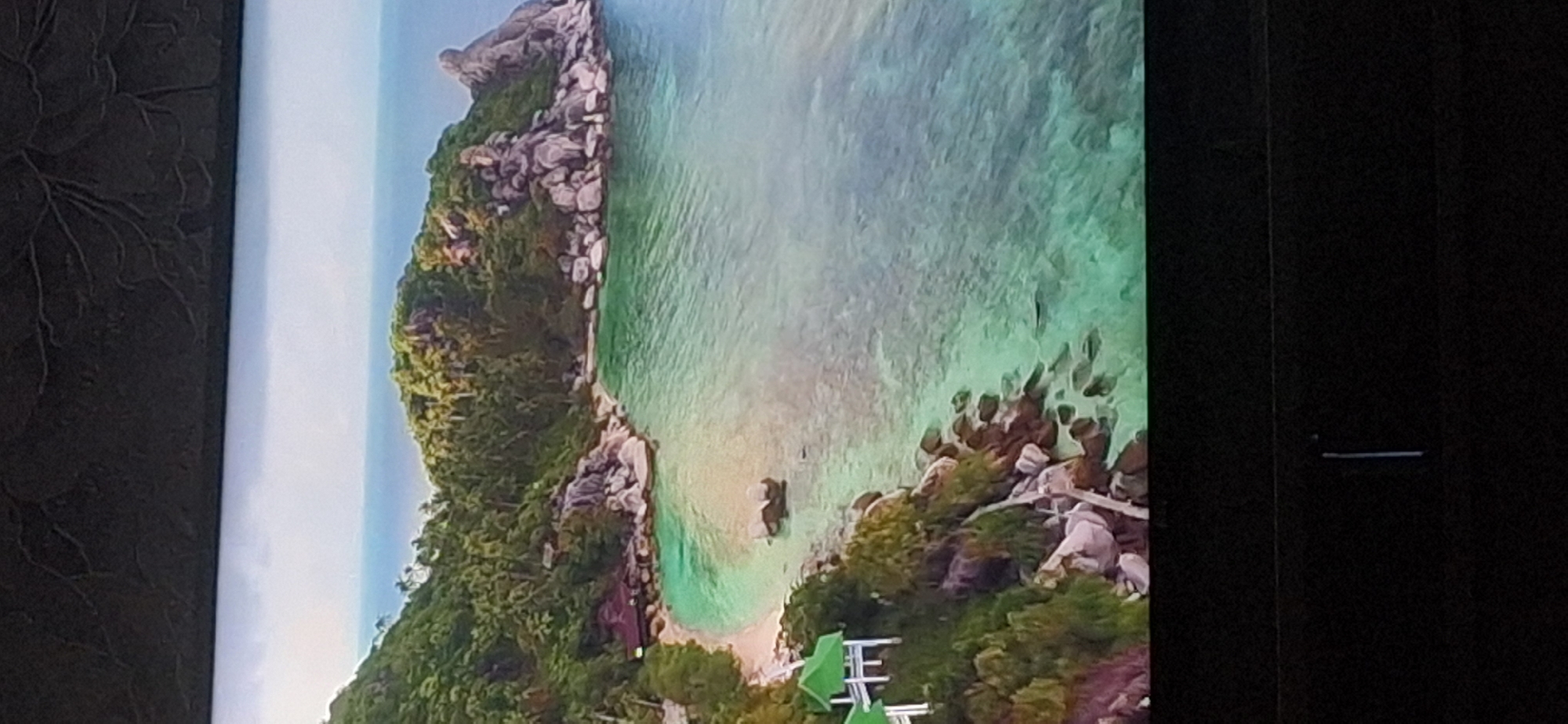 This is an aerial, wide-angle shot of a tropical island cove. Lush green foliage covers the steep hillsides that frame a crescent-shaped, sandy beach. The water is a clear, turquoise color, transitioning to deeper blue in the distance. Large, grey rocks are scattered along the coastline and in the shallow water. There are several small, traditional-style buildings with green roofs and white structures visible on the left side of the image, nestled amongst the trees. A single, dark-colored boat is anchored in the shallow water off the beach. The sky is overcast and a pale grey. The foreground of the image is dark and out of focus, suggesting it might be the edge of a screen or viewing device. There is no indication of the city of Bila Tserkva, Ukraine, or any urban environment in this image.