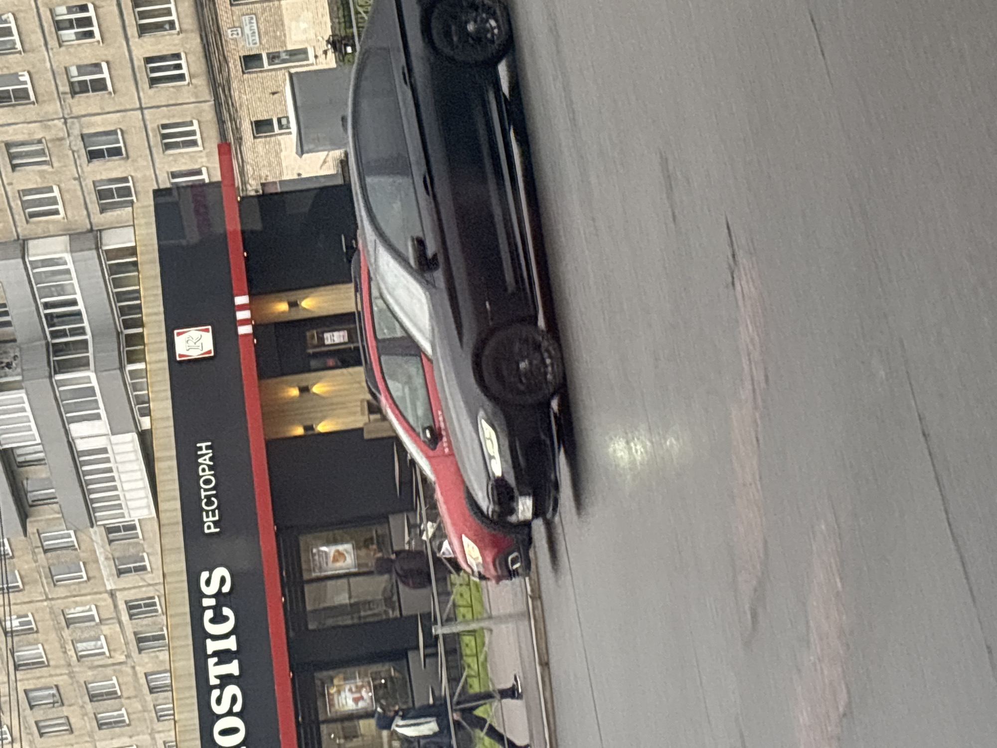 The image shows a street scene in what is identified as Unknown, Russia, with a building that houses a restaurant called "KOSTIC'S." The sign outside the restaurant also includes the word "PECTOPAH," which is "restaurant" in Russian. Adjacent to the restaurant is a multistory residential building. A black and red vehicle is parked on the street beside the restaurant. A pedestrian is visible walking on the sidewalk. The street appears to be paved.
