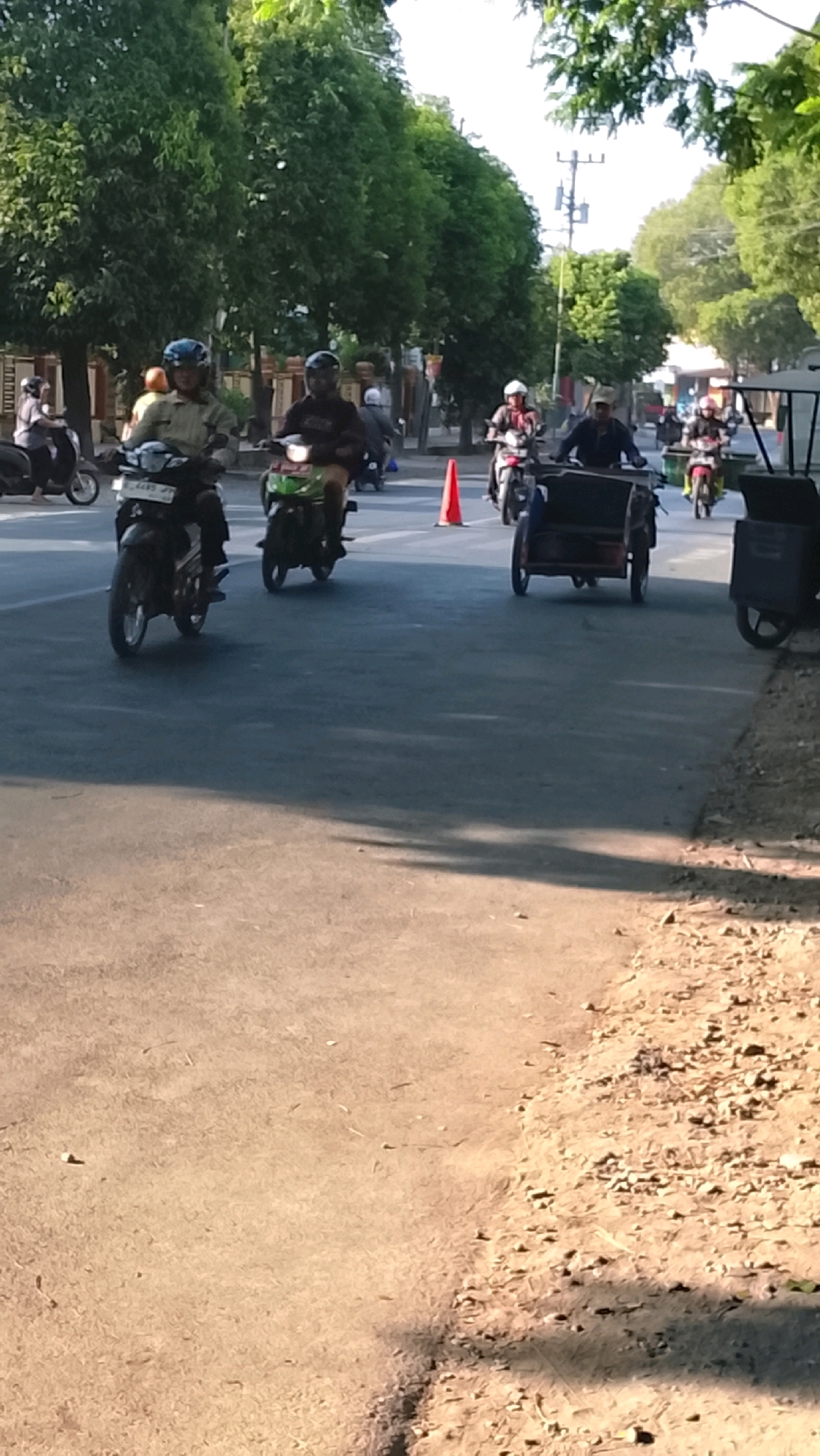 The image depicts a street scene in Slawi, Indonesia, with several people riding motorcycles. In the foreground, a black motorcycle is parked on the right side of the road. Further down the street, multiple motorcycles are in motion. On the left, a person wearing a light-colored shirt and a blue helmet rides a black motorcycle. Next to them, another person in a dark shirt and helmet rides a green motorcycle. In the middle distance, a person in a red and white checkered shirt on a motorcycle is visible, followed by a three-wheeled vehicle. On the far right, a black storage container is parked. The road is paved, with a sandy shoulder on the right side. Trees line the left side of the street, and power lines are visible in the background. The scene appears to be during the daytime, with sunlight casting shadows.