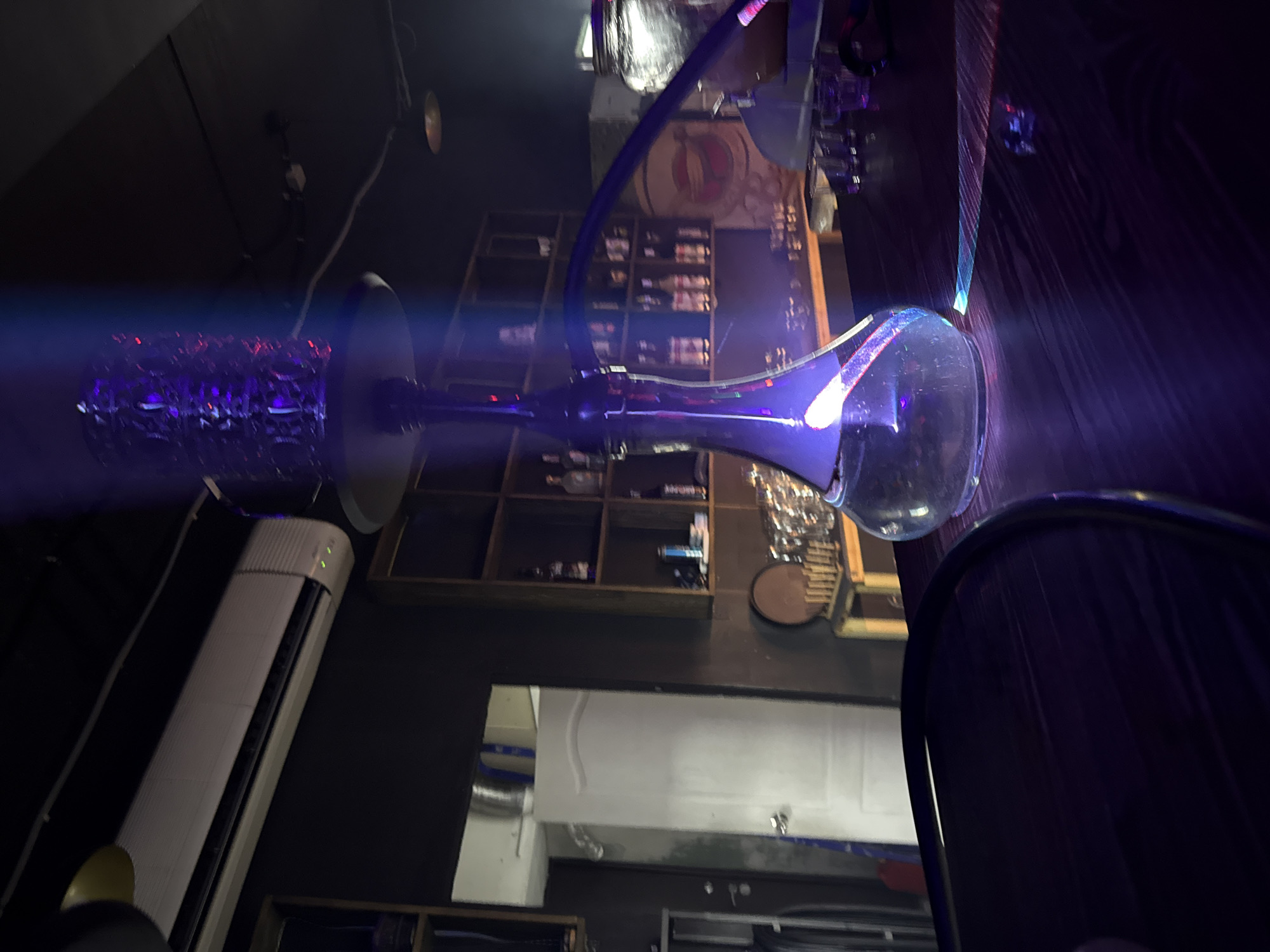 The rotated image captures a dimly lit indoor scene, possibly a lounge or bar in Tobolsk, Russia, characterized by atmospheric purple lighting. The central subject is a modern hookah, with a transparent, bell-shaped glass base positioned towards the right side of the image. Its dark, possibly metallic, stem extends towards the left. A prominent, bright purple light emanates from the hookah, creating a vivid, horizontally extending beam that pierces the dark ambiance and highlights a textured, reflective component on the hookah's upper section.
In the upper left quadrant of the image, a long, white, wall-mounted air conditioning unit or heater is visible against a dark wall. Below the hookah, towards the bottom of the image, portions of a dark wooden surface, likely a table, are visible. The background, largely to the top and left of the hookah, features dark wooden shelving units stocked with various bottles, suggesting an establishment serving drinks. Above these shelves, a circular emblem or logo is faintly visible on the wall, though its details are indistinct. Further in the background, near the top, a silver corrugated air duct can be seen.
The scene conveys an evening or nighttime setting, emphasized by the artificial purple illumination and dark surroundings. No individuals are present in the image, and no clear text is visible on any objects or surfaces. The overall mood is cool and contemporary, typical of a hookah lounge or nightclub setting.