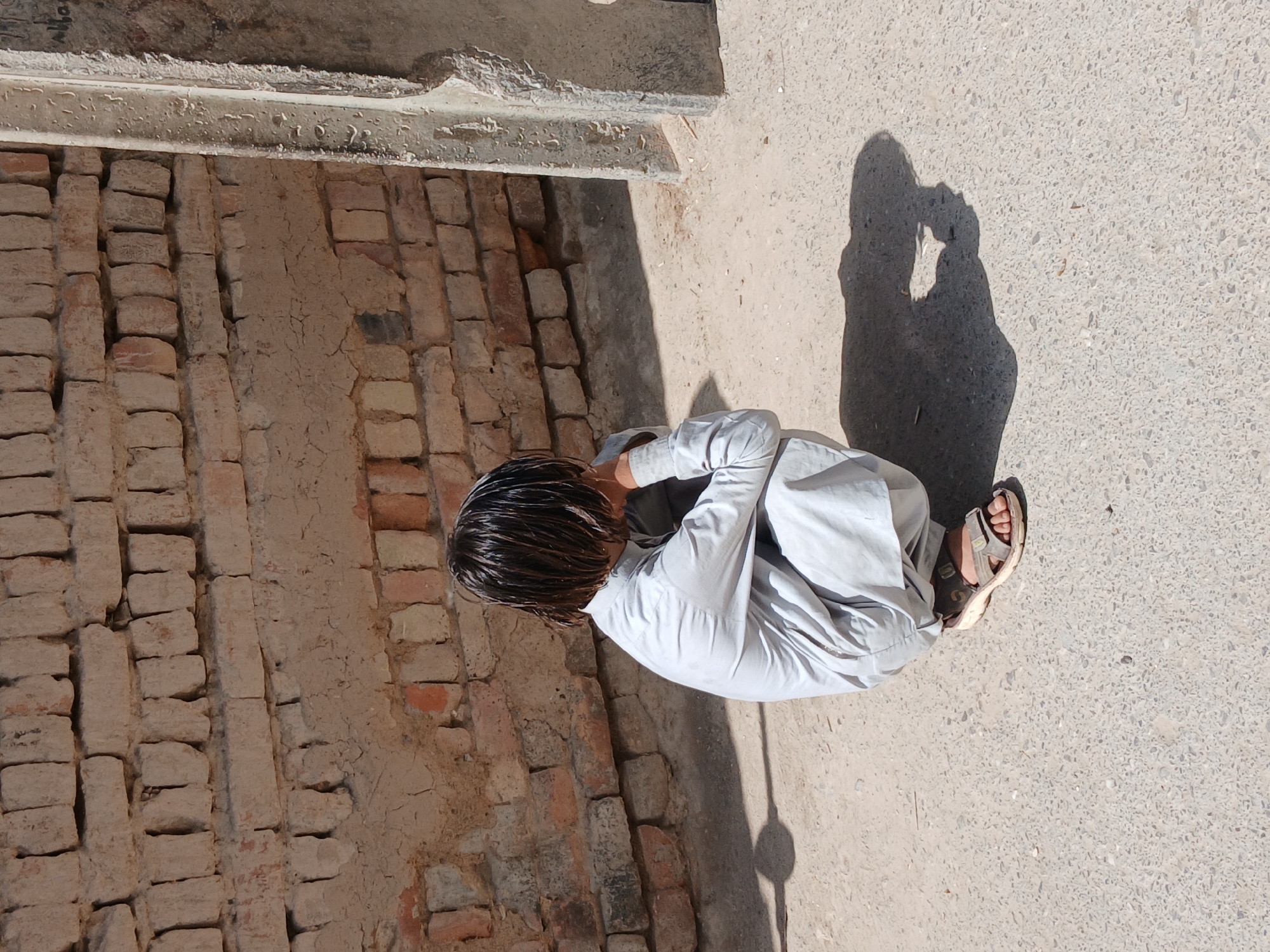 A person sits cross-legged on a light-colored, speckled concrete surface, with a brick wall to their left. The person has dark, wet hair and wears a light-colored, long-sleeved garment and sandals. The person casts a shadow on the concrete. A portion of a building or structure overhangs the scene at the top of the frame. The location is the city of Quetta City Tehsil, Pakistan.