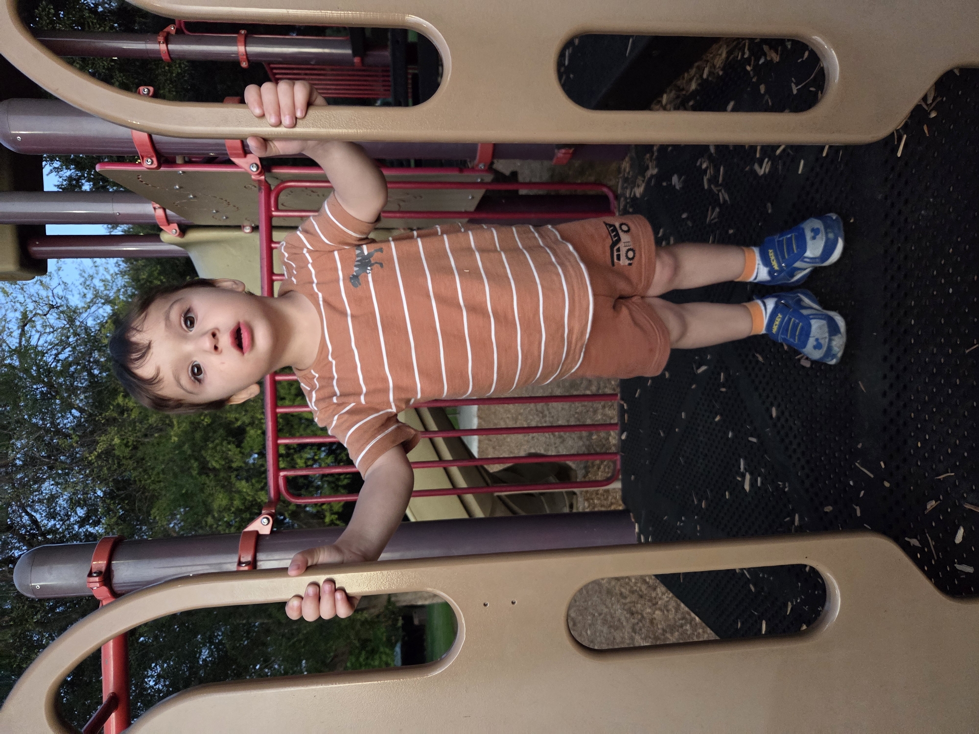 A young boy stands on a playground structure, holding onto the plastic supports on either side. He is wearing an orange and white striped t-shirt with a dinosaur graphic and matching orange shorts with a truck graphic. His feet are clad in blue and white sneakers with "MICKEY" written on them, and white socks with orange trim. The playground features a dark grey rubber matting floor and a red metal railing in the background. Trees are visible behind the structure. The lighting suggests it is late afternoon or early evening. The image was taken in Glendale, United States of America.
