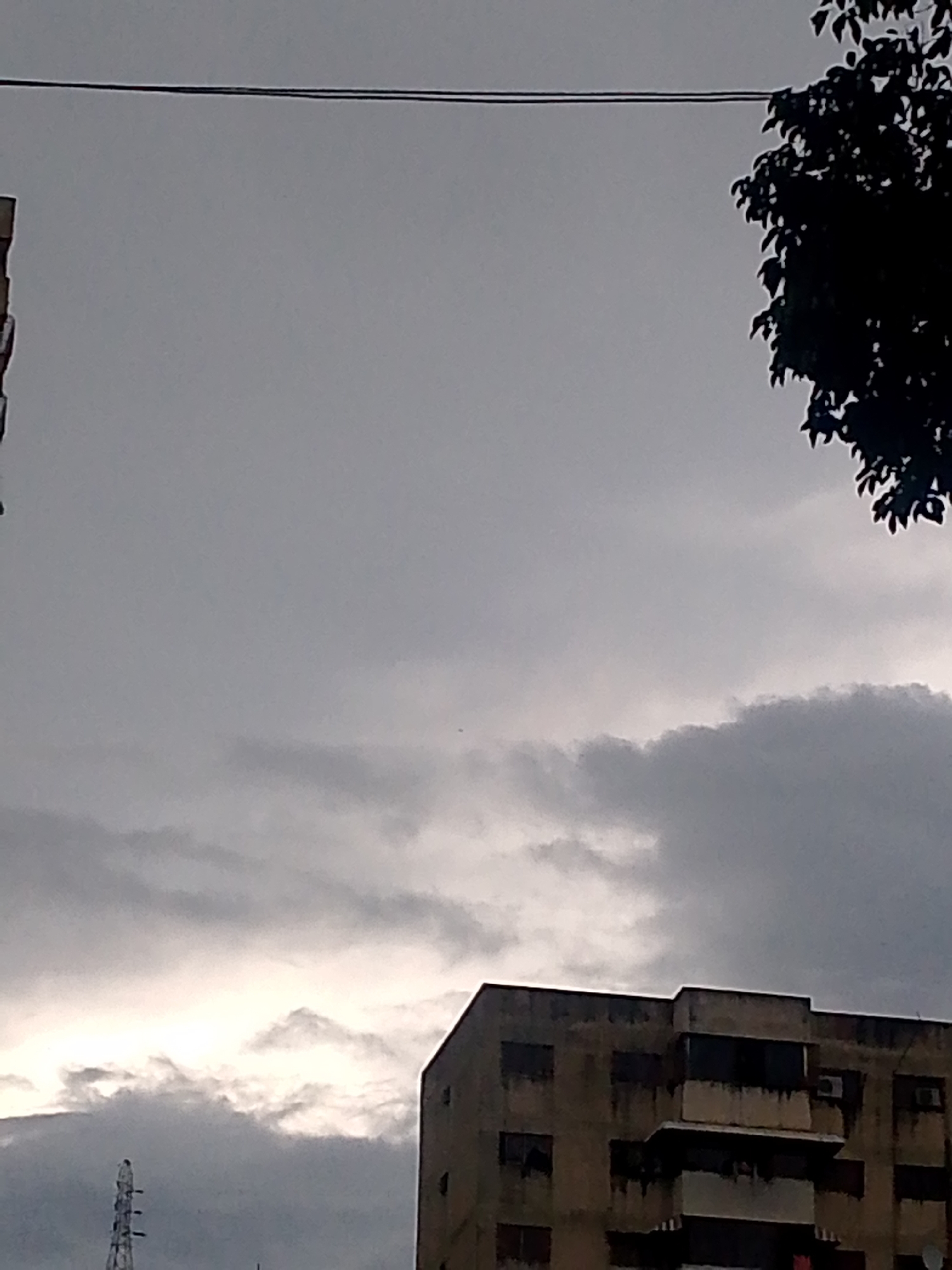 The image captures a cloudy sky over a city, likely Caracas, Venezuela, with overcast conditions dominating the scene. In the foreground, a portion of a weathered, multi-story apartment building is visible on the right side, its facade appearing stained and aged. A balcony with a railing and a darker recessed area is prominent. To the left of this building, a transmission tower with a lattice structure stands against the sky. The sky itself is filled with a mix of dark and lighter gray clouds, with a bright, luminous band suggesting the presence of the sun behind thicker cloud cover. On the far right, the silhouette of tree branches is visible against the sky, partially obscuring the view. A thin, dark wire stretches horizontally across the upper part of the frame, potentially a power line or communication cable. The overall impression is one of a typical urban environment under a heavy sky.