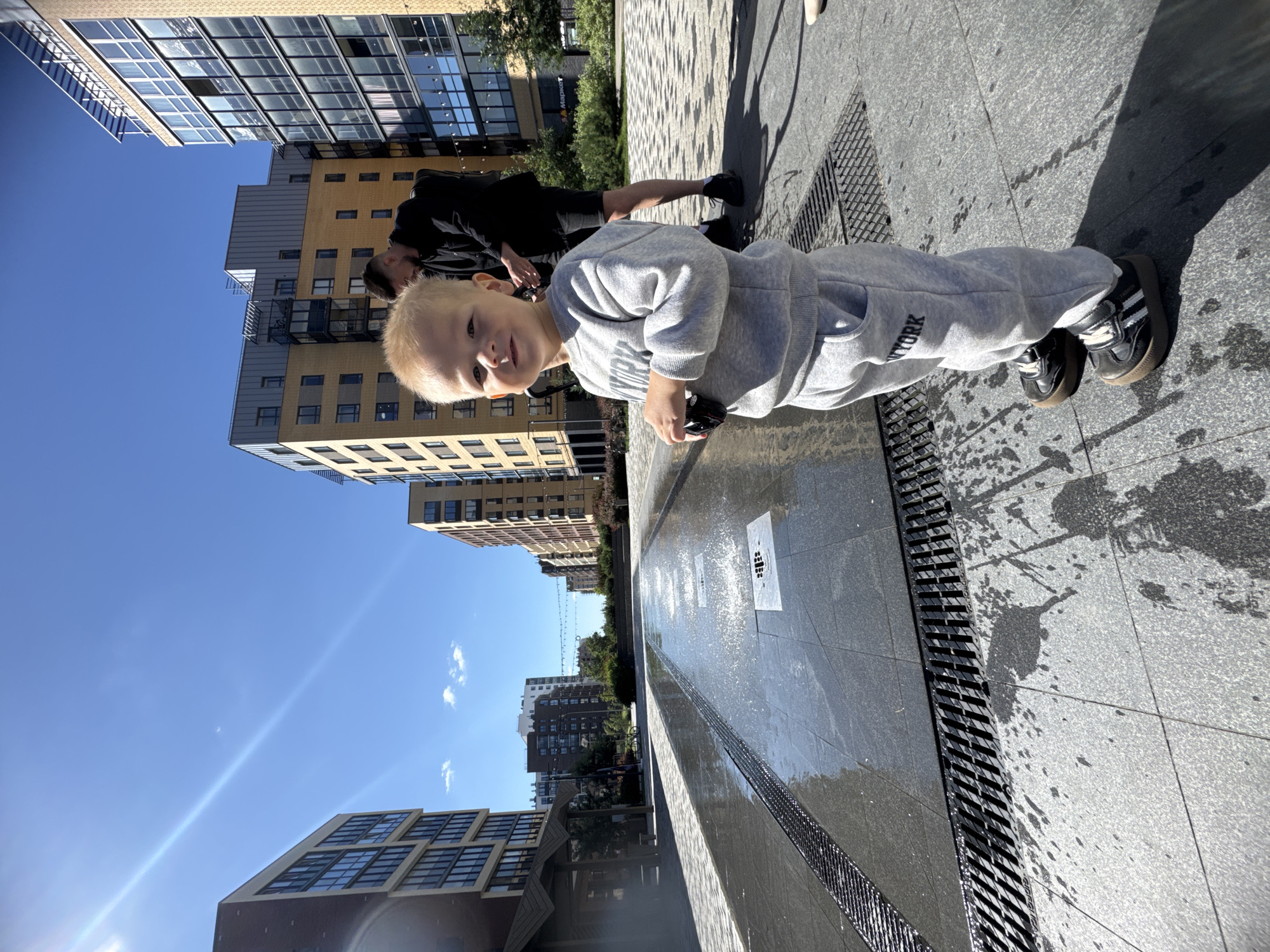 A young child with blond hair and a light grey tracksuit stands in the foreground, facing the camera with a slight smile. The child's left hand is holding a dark object. Behind the child, a man in a black t-shirt and shorts is visible, with his back to the camera, looking down. They are standing on a paved area with water jets spraying from the ground, creating a shallow water feature. In the background, modern apartment buildings with numerous windows rise against a clear blue sky. The location is identified as Novoselye, Russia. The environment appears to be a public urban space on a sunny day.