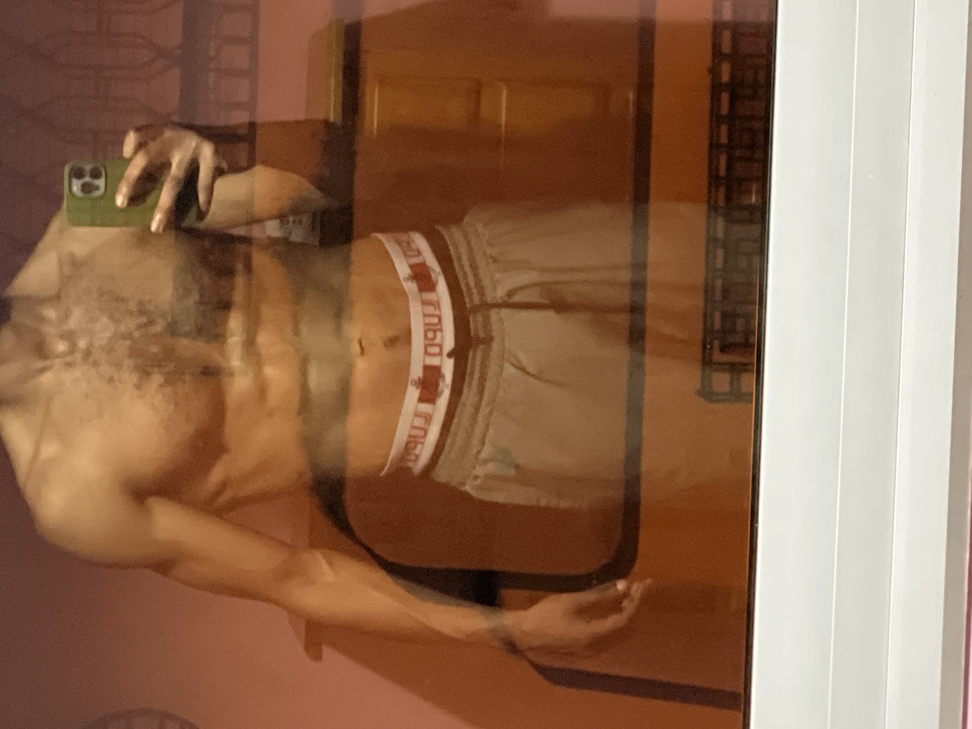 A person with a muscular physique is taking a mirror selfie with a phone in their left hand. They are wearing light-colored shorts with a waistband featuring a repeated logo. The reflection shows a partially visible room with furniture and light-colored walls. The location is likely indoors, potentially in M'Diq, Morocco. The photograph is taken at night.