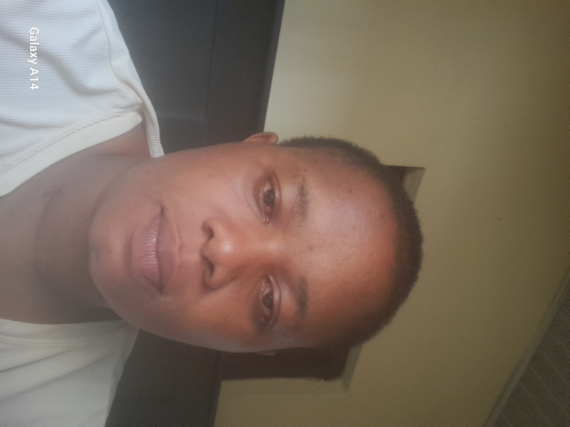 A close-up, slightly angled shot of a woman's face, possibly a selfie, taken indoors. The woman has short, dark hair and appears to be wearing a light-colored, ribbed top. The background consists of a dark, undefined area and a light yellow wall. The location, Udi, Nigeria, is not visually confirmed in the image. The woman is looking at the camera. Text "Galaxy A14" is visible in the upper left corner.