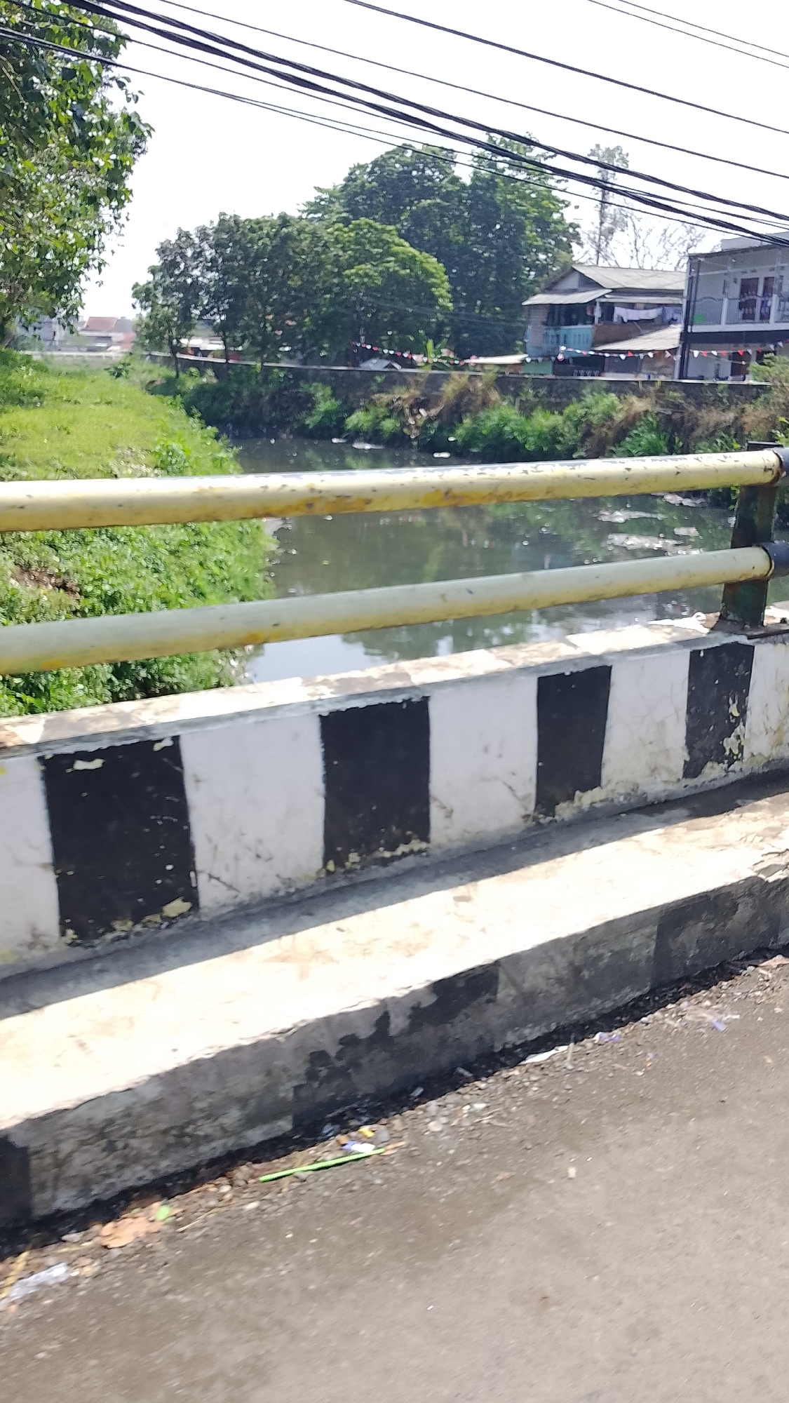 A sunlit scene captures a bridge railing overlooking a narrow waterway lined with green foliage and trees. Beyond the waterway, a residential area is visible with houses and a glimpse of a building with multiple levels. Power lines are strung across the top of the frame. The bridge railing is painted yellow, and the concrete barrier below it is marked with alternating black and white stripes. Debris is present on the road surface near the edge of the bridge. The overall impression is one of a semi-urban or suburban environment in Bojongsoang, Indonesia.