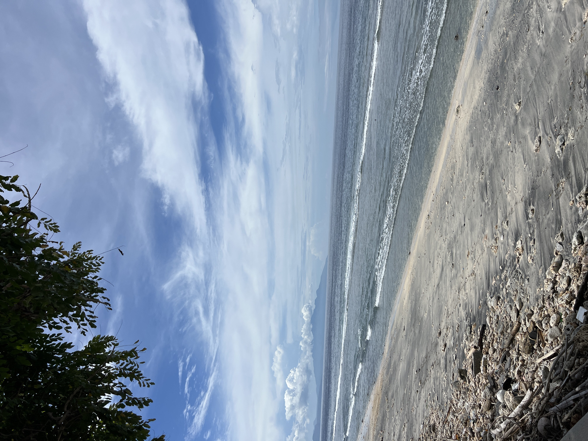 The image depicts a beach scene in Nusa Penida, Indonesia. The sky is partly cloudy, with a mix of blue sky and white, feathery clouds. The sea is calm with small waves lapping the shore. The beach itself is a mix of dark sand and an area littered with pebbles, shells, and debris. Vegetation is visible in the lower left corner of the image, with leaves and branches suggesting a tree or shrub. The image is taken from a slightly elevated perspective. There are no people visible in the scene.