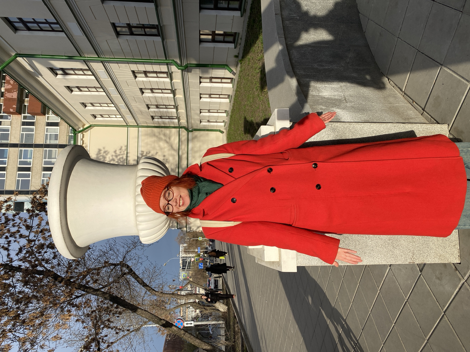 A smiling young woman is the central subject of this sunlit outdoor photograph, taken in an urban setting identified as Yekaterinburg, Russia. She wears a vibrant, long red coat, a green scarf, and an orange knitted beanie, along with glasses. Her expression is joyful, with a wide smile directed towards the camera, and her arms are slightly outstretched in a relaxed pose.
She is standing next to a very large, white, stylized chess piece, which appears to be either a king or queen due to its shape and prominent size. The woman is positioned on a paved area, possibly a sidewalk or plaza, with a low stone barrier or bench to her right.
The background features several multi-story buildings. A notable one has a light-colored facade with numerous rectangular windows and distinctive green piping along its exterior. Bare-branched trees are present in the foreground and background, suggesting the season is late autumn, winter, or early spring. In the distance, a street is visible with cars and other pedestrians. The sky is clear and blue, and the bright sunlight casts pronounced shadows on the ground, indicating either morning or late afternoon. Street signs are visible in the background, but any text on them is too distant to be legible, although traffic symbols are discernible. The overall atmosphere is cheerful and bright.