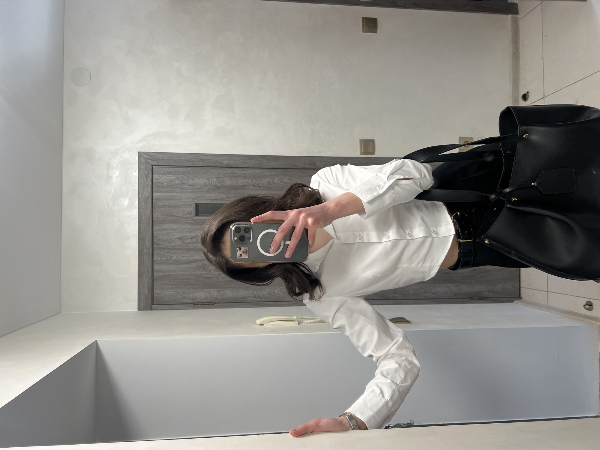 A woman is taking a mirror selfie in what appears to be a hallway or entrance area of an apartment building in Ternopil, Ukraine. She is wearing a white, cropped button-up shirt and black pants with a black belt. She is holding a black tote bag in her right arm and her left hand is resting on a white wall to her left. Her phone, with a case featuring a cat sticker, is visible as she takes the photo. The background includes a gray wooden door, a beige wall with a textured finish, and a tiled floor. There is also a light-colored intercom system mounted on the wall.