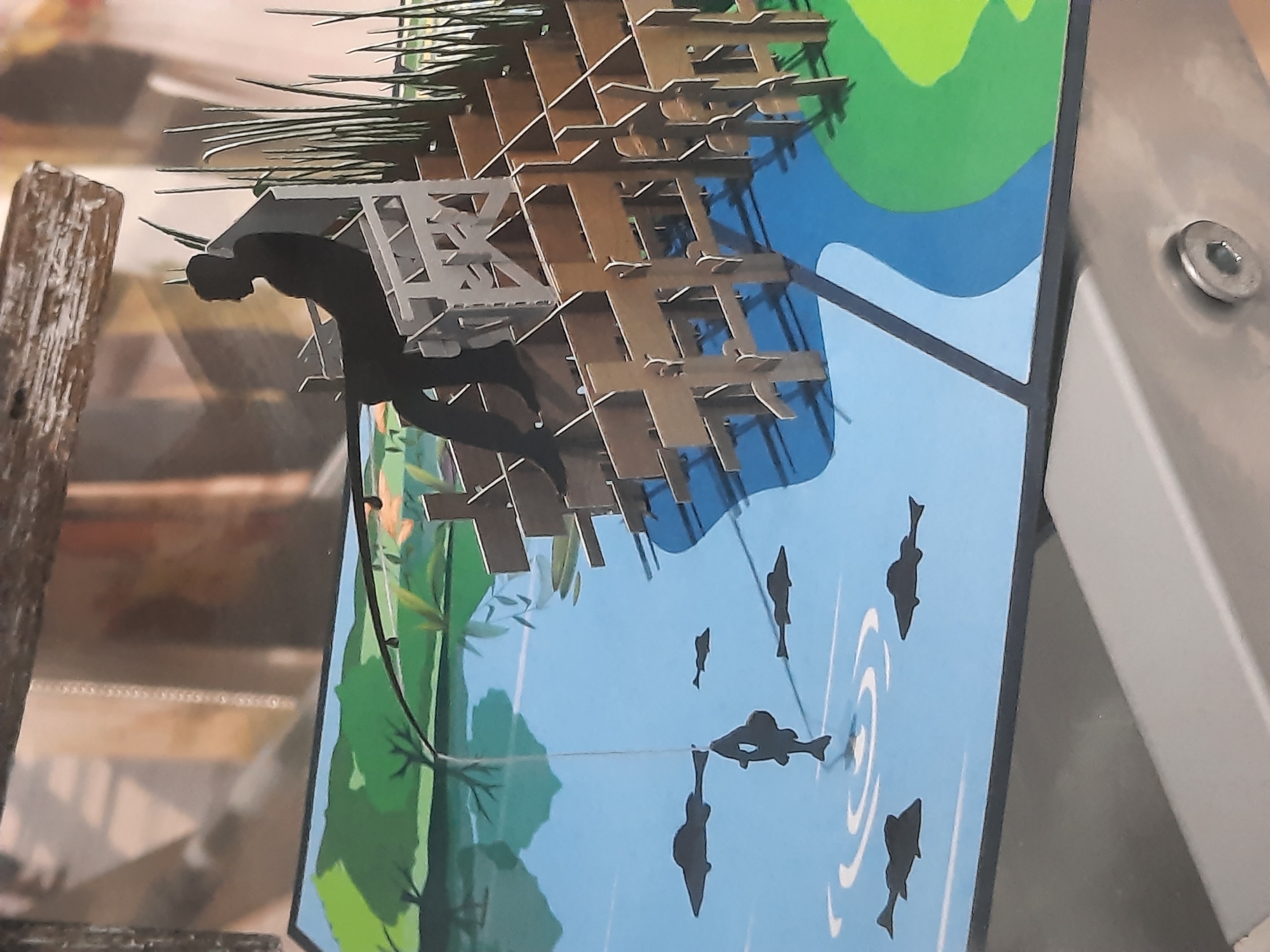 The image shows a pop-up card depicting a silhouette of a person fishing from a chair on a wooden structure. The fishing line extends into a blue body of water where several black fish silhouettes are visible. The background features green land with trees and tall grass. The card is placed on a reflective surface, possibly a table, with a hint of a screw in the foreground. The scene is a stylized representation of an outdoor activity. There is no indication of the city of Rosenheim, Germany, within the image.