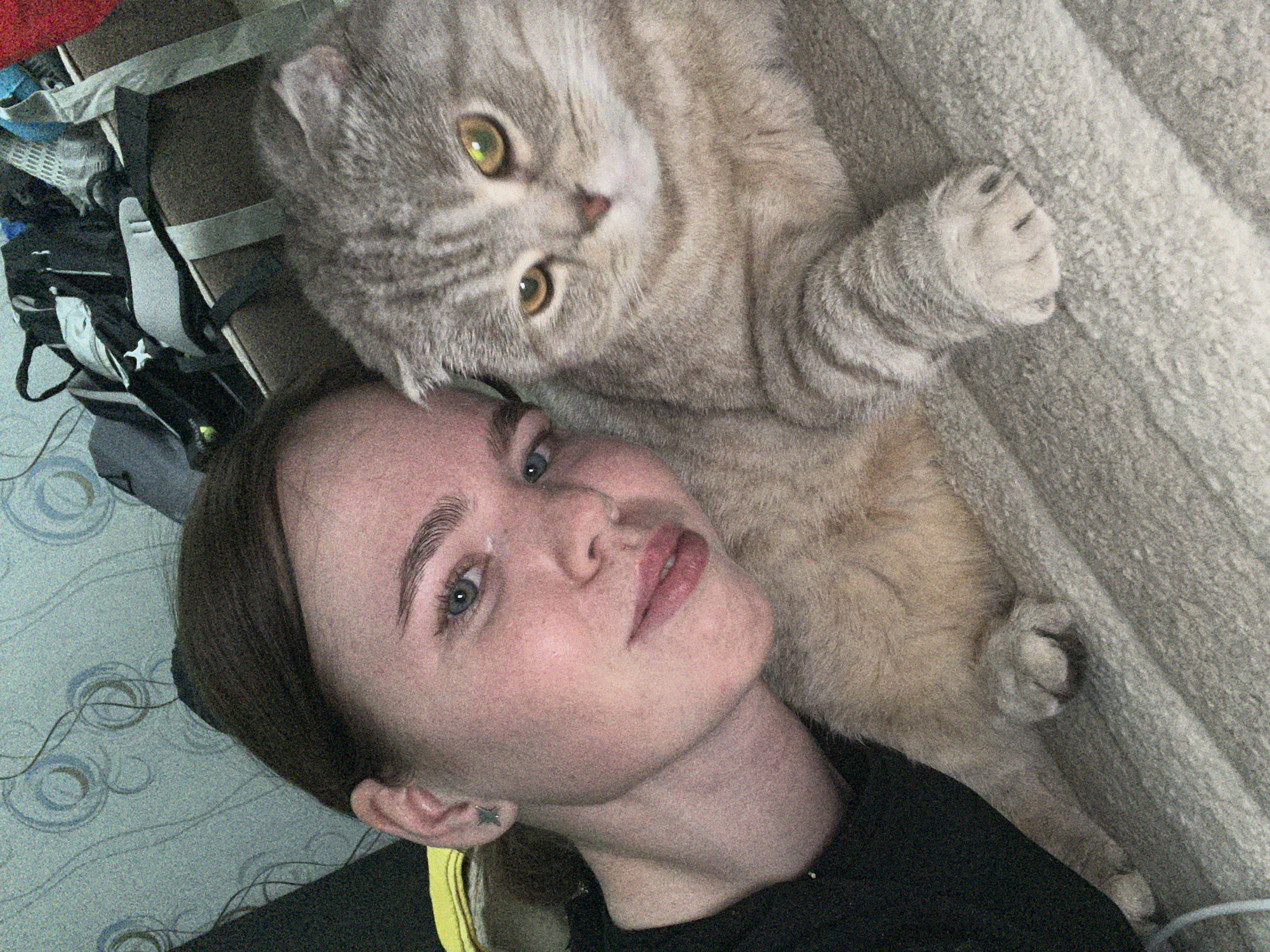 A young adult female and a domestic cat are depicted in a close-up indoor portrait. The female, positioned on the left side of the frame, has dark brown hair, light-colored eyes, and a subtle smile, wearing a dark-colored top. A small, star-shaped metallic earring is visible in her left earlobe. To her right is a Scottish Fold cat, characterized by its folded ears, grey and white tabby patterned fur, and prominent yellow-green eyes. The cat is looking directly at the camera with a neutral expression, resting with one paw visible on a textured surface. Both subjects are situated on a light-grey, textured blanket or bedspread. The background on the left features wallpaper with a repeating pattern of faint blue and grey concentric circles. The upper right portion of the background shows a cluttered area containing a dark-colored backpack with light accents and other indistinct items. The image exhibits a moderate level of digital noise. No specific location within the city of Chaikovsky, Russia, is discernible from the visual information provided.