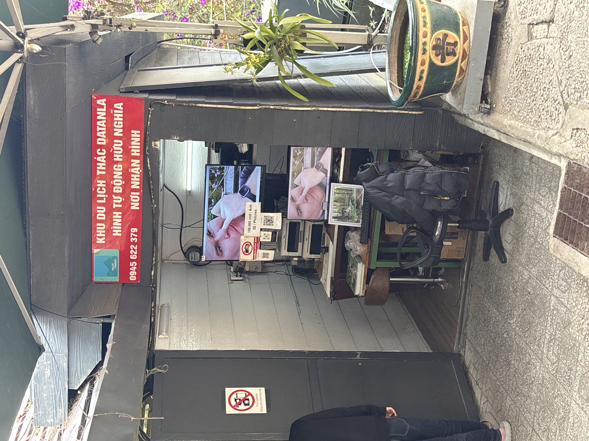 The image shows an outdoor booth or small shop, likely serving as an information or reception point for a tourist attraction in Da Lat, Vietnam. The booth has a dark, possibly wooden, exterior.
A prominent red vertical sign is affixed to the left side of the booth. Inside, two screens are visible, displaying close-up images of a person's face. Below these screens, a counter holds various items, and a black jacket hangs on a chair. The floor inside the booth appears to be made of light-colored planks.
Above the booth, a lush green potted plant with small yellow flowers hangs. To the right, a large green ceramic pot decorated with a gold-colored Chinese character "贵" (gui, possibly meaning noble or expensive) rests on a concrete ledge. The surrounding area features textured light-colored pavers on the ground and a rough concrete wall.
A person, visible from the waist down, stands in front of the booth's entrance, dressed in dark clothing and white shoes. The scene appears to be set during the daytime, possibly under shaded or overcast conditions.
Visible text on the red sign reads: "KHU DU LỊCH THÁC DATANLA" (Datanla Waterfall Tourist Area), "HÌNH TỰ ĐỘNG HỮU NGHĨA" (Automatic/Self-service Photos Friendly), "NƠI NHẬN HÌNH" (Photo Reception Place), and a phone number "0945 622 379". A "DALAT TOURIST" logo is also present. Another sign on the lower part of the booth displays a "No Camera" symbol along with the Vietnamese text "Không quay phim chụp ảnh" (Do not film or take photos). A price list is also visible, stating "100.000 vnd/ Ảnh" (100,000 VND / Photo) and "5$/Pictures" (5 USD / Pictures).