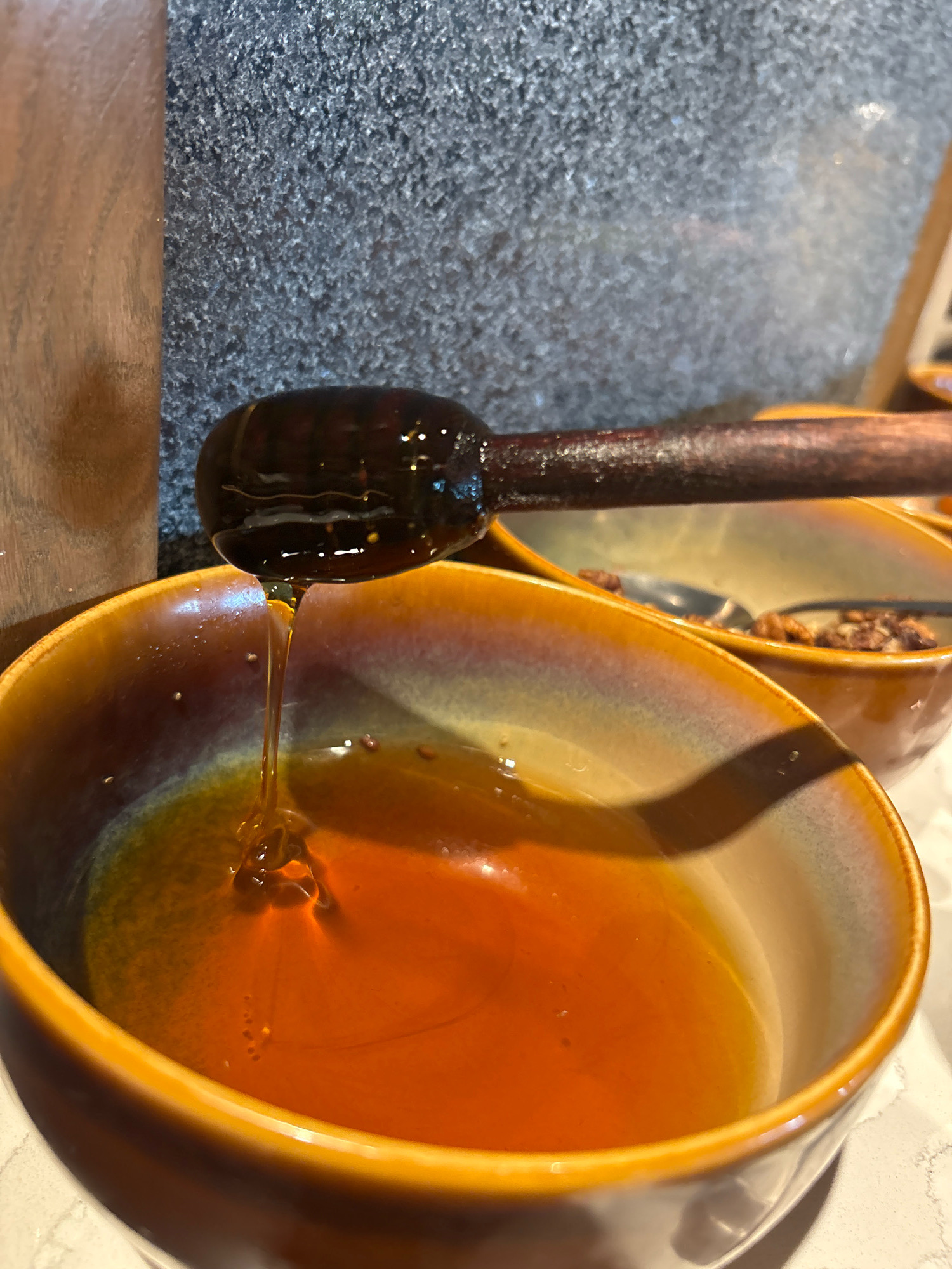 In Vagator, India, a wooden honey dipper with honey dripping from it hovers over a ceramic bowl filled with amber-colored honey. The bowl has a gradient glaze transitioning from a light beige at the rim to a deep orange-brown towards the center. Behind the honey bowl, another bowl is visible, containing nuts and a spoon. The background consists of a textured gray surface and a wooden panel, suggesting a rustic or natural setting.