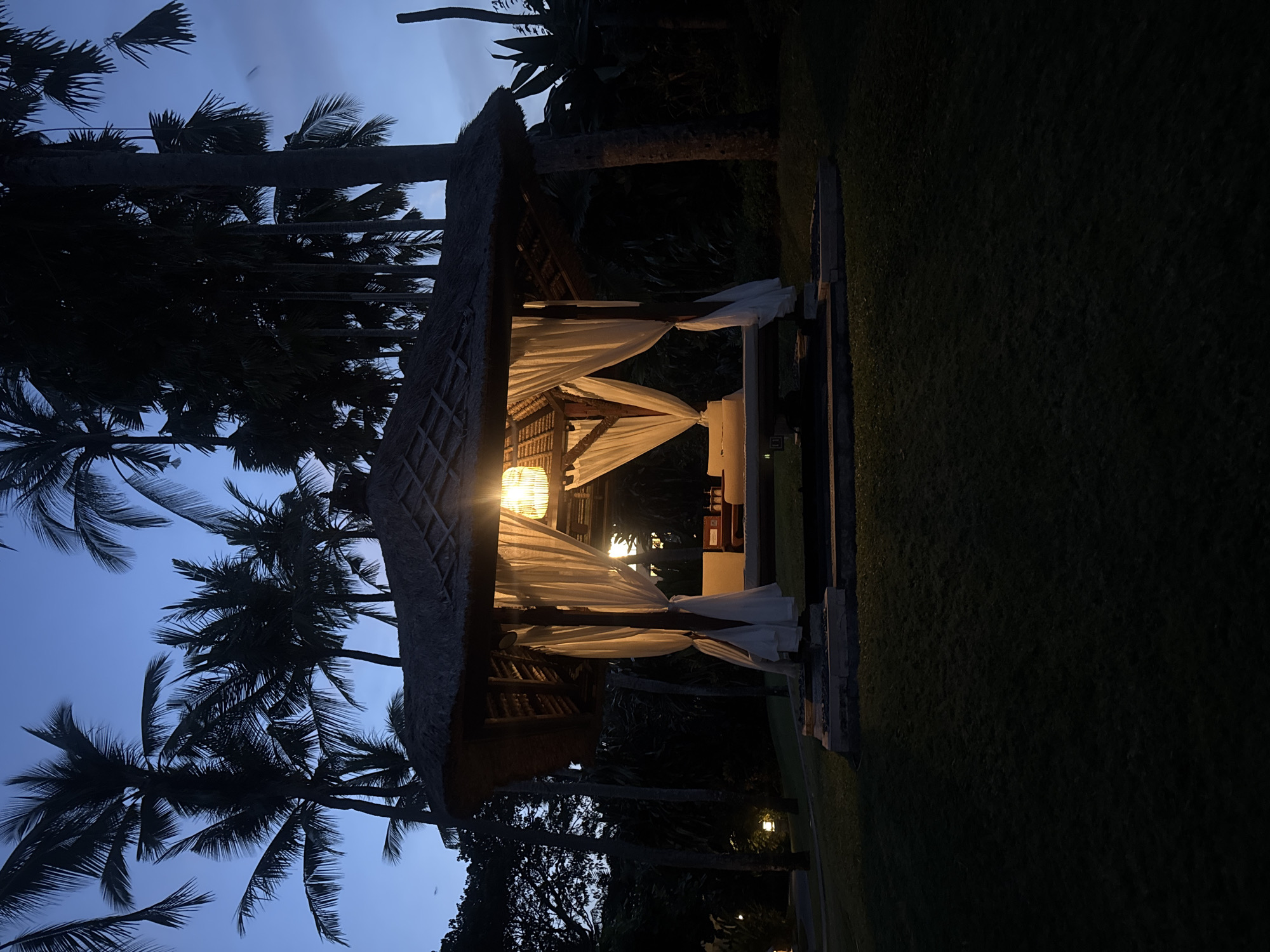 The image displays an illuminated outdoor gazebo at dusk or night, located in Nusa Dua, Indonesia. The central subject is a hexagonal or octagonal gazebo with a distinct thatched roof and exposed wooden support beams. White, opaque drapes are tied back to the interior wooden posts, partially enclosing the structure. A single, ornate lantern with a warm, yellowish glow hangs from the center of the gazebo's ceiling, providing the primary illumination for the interior. Inside, an elevated platform, likely a daybed or massage table, is visible, accompanied by what appear to be pillows or cushions. Several small bottles are arranged on a surface within the gazebo to the right. The gazebo is situated on a raised, multi-tiered stone base on a manicured grass lawn, which appears dark green due to the low light. In the background and extending upward, numerous tall palm trees and dense tropical foliage are silhouetted against a deep blue twilight sky. A faint ambient light source is visible on the far left of the image, illuminating a small section of the background. No individuals or specific actions are depicted.