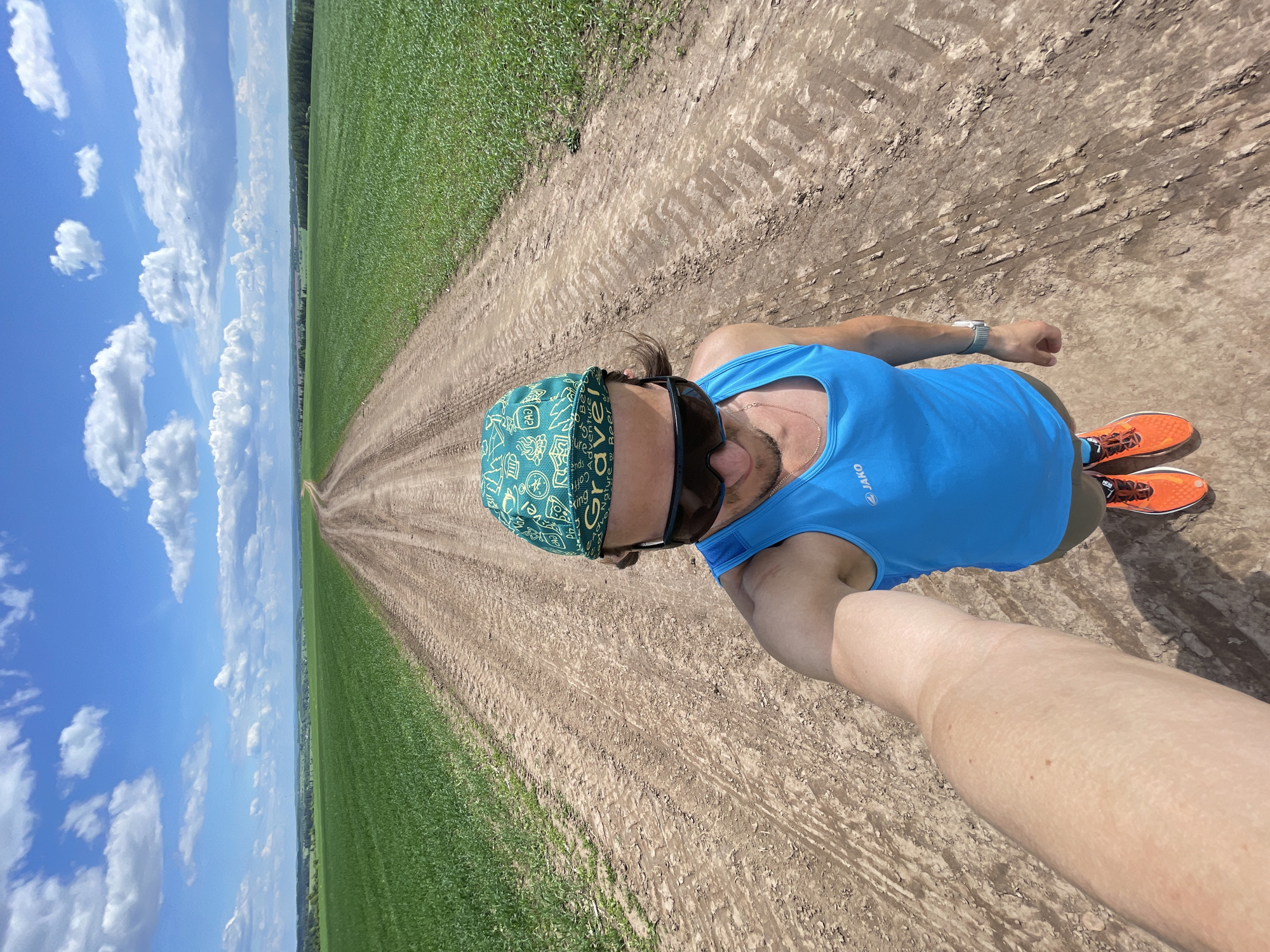 A low-angle selfie captures a male individual standing centrally on a dirt road, looking downwards towards the camera, which is held by an extended right arm. The individual wears a green and white patterned bandanna with "Gravel" text and adventure-themed graphics, dark wraparound sunglasses, a light blue sleeveless athletic tank top with a white "JAKO" logo on the left chest, a thin gold chain, and bright orange athletic shoes with black laces. A silver/grey smartwatch is visible on the left wrist. The dirt road, marked with tire tracks, extends into the distance, flanked on both sides by vast, vibrant green fields of crops. The horizon reveals a distant treeline and a subtly undulating landscape under a bright blue sky dotted with scattered white cumulus clouds. No specific city or urban structures are visible; the location is Unknown, Unknown.