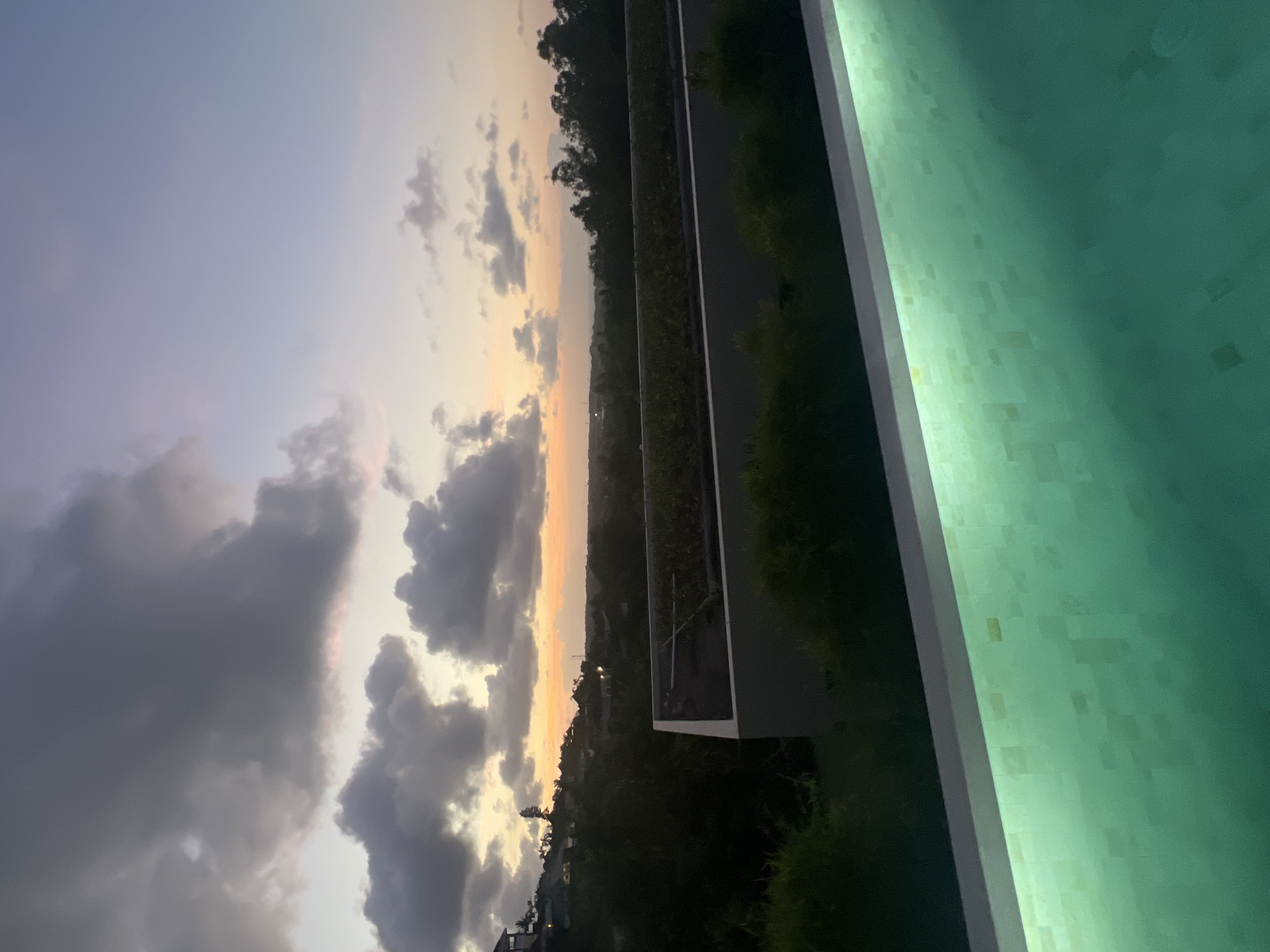 The image depicts an outdoor scene in Kuta Selatan, Indonesia, captured during twilight or sunset. A sky filled with dark, gray clouds transitions to lighter hues towards the horizon, where an orange glow suggests the setting sun. Below the horizon, a vague cityscape is discernible, possibly residential areas with scattered lights.
A dark, linear feature cuts through the landscape, likely a road or waterway bordered by vegetation. In the foreground, a large, blurry, light green surface dominates the right side, possibly a building exterior or a pool with tiled finish, brightly lit. The view appears to be taken from an elevated position, possibly a balcony or higher floor of a building.
