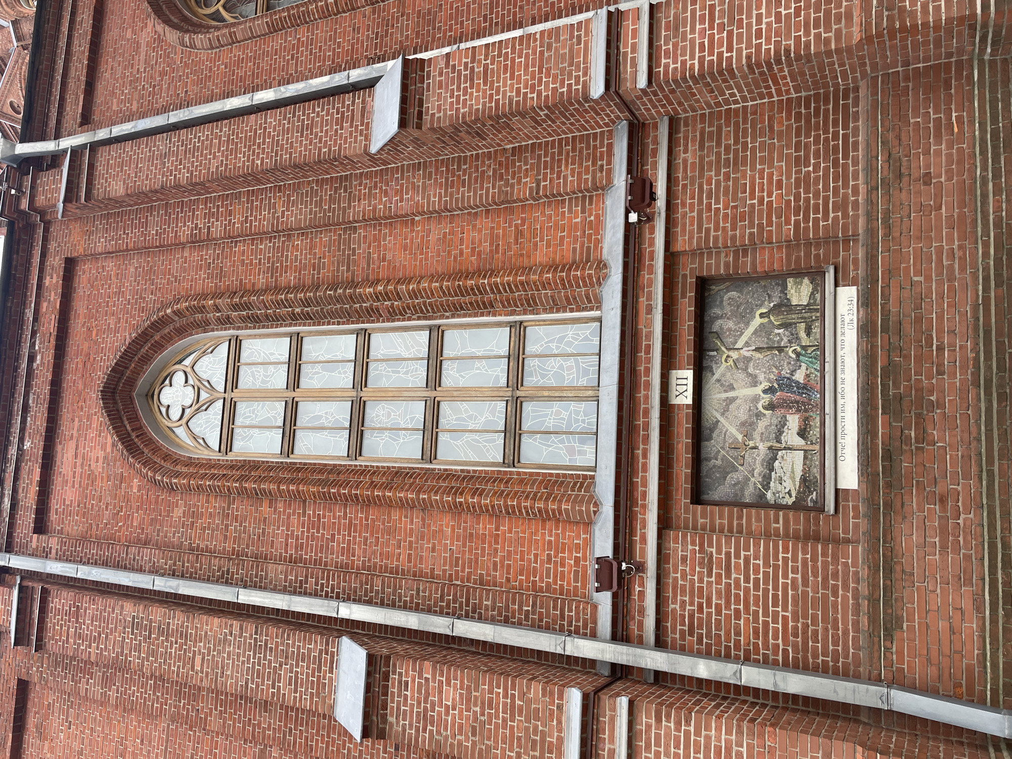 The image shows the brick facade of a religious building in Moscow, Russia. A tall, arched stained-glass window is centrally located, divided into multiple panes by wooden mullions. Below the window, a rectangular mosaic depicts a scene with three figures, the central one on a cross, suggesting a crucifixion. To the right of the mosaic, the Roman numeral "XII" is visible. Beneath the mosaic, a white plaque bears text in Cyrillic script and a biblical reference. A metal downspout runs vertically along the left side of the building, and there are decorative brickwork elements and projecting ledges. The overall impression is of a historical and religious structure with intricate architectural details.