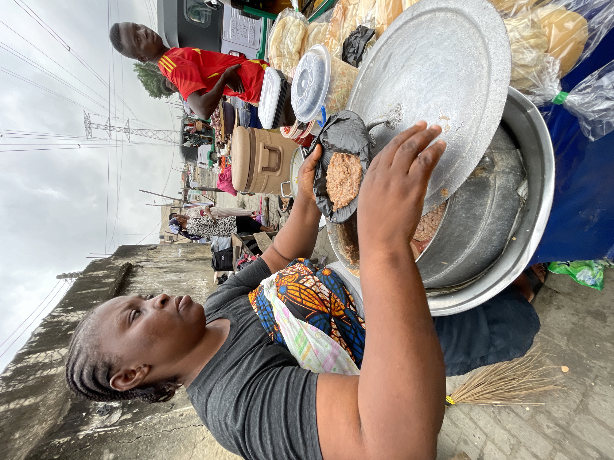 This image captures a street food vendor in Ado, Nigeria. The vendor, a woman with braided hair, is wearing a dark grey t-shirt and a vibrantly patterned fabric wrapped around her waist. She is in the process of serving food from a large, metallic pot with a silver lid, which she is holding open with her left hand. In her right hand, she holds a black disposable plate filled with a reddish-brown, grainy food.
To the right of the main vendor, a young man wearing a red t-shirt with yellow stripes is visible, looking towards the camera. Behind them, other vendors and customers are present, along with various food items such as bread rolls, containers of coleslaw, and what appear to be onions in a large bowl. Several vehicles, including a tricycle and a larger green and white vehicle, are parked nearby, suggesting a busy marketplace or roadside setting. The background shows a concrete wall and overhead power lines, characteristic of an urban environment. The sky is overcast, indicating a cloudy day.