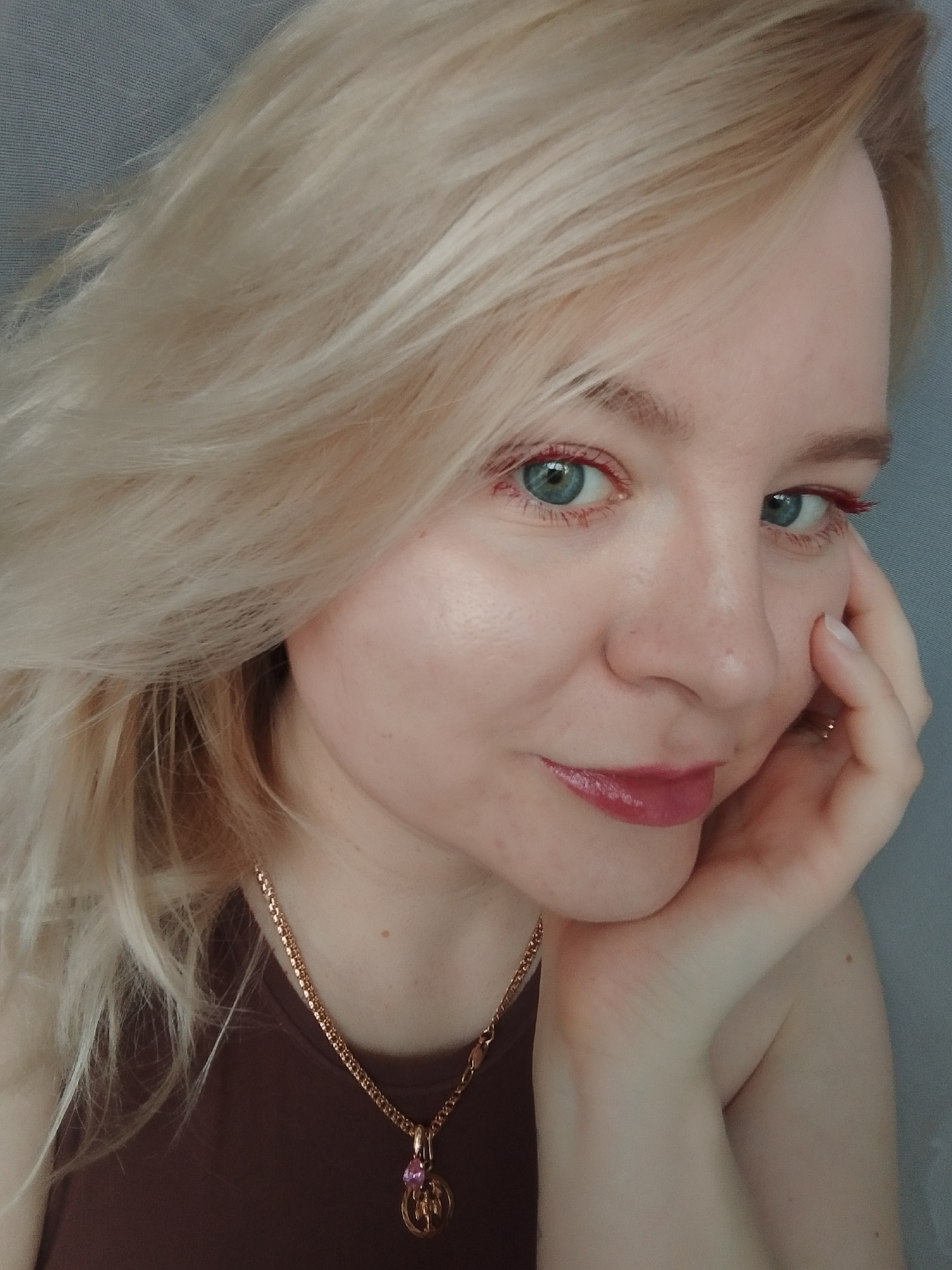 A close-up portrait of a fair-skinned woman with blonde hair and blue eyes. She is looking directly at the camera with a slight smile. She is wearing a brown sleeveless top and a gold necklace with a pendant. She is touching her face with her right hand. The background is a plain gray color. There is no evidence that this image was taken in Izhevsk, Russia.
