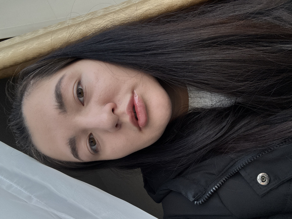 The image displays an upward-angled, medium close-up self-portrait of an adult female. She has long, dark brown hair falling over her shoulders, brown eyes, and full, dark eyebrows. Her lips are slightly parted and appear glossy. She wears a black zipped jacket with a silver snap button visible on the left chest area, over a light grey knitted sweater. The subject gazes directly into the camera. The background consists of a translucent white curtain on the left side and a textured, light brown curtain on the right, indicating an indoor setting adjacent to a window. The image provides no specific visual information to identify the location as Kurovskoye, Russia, or any external environment.