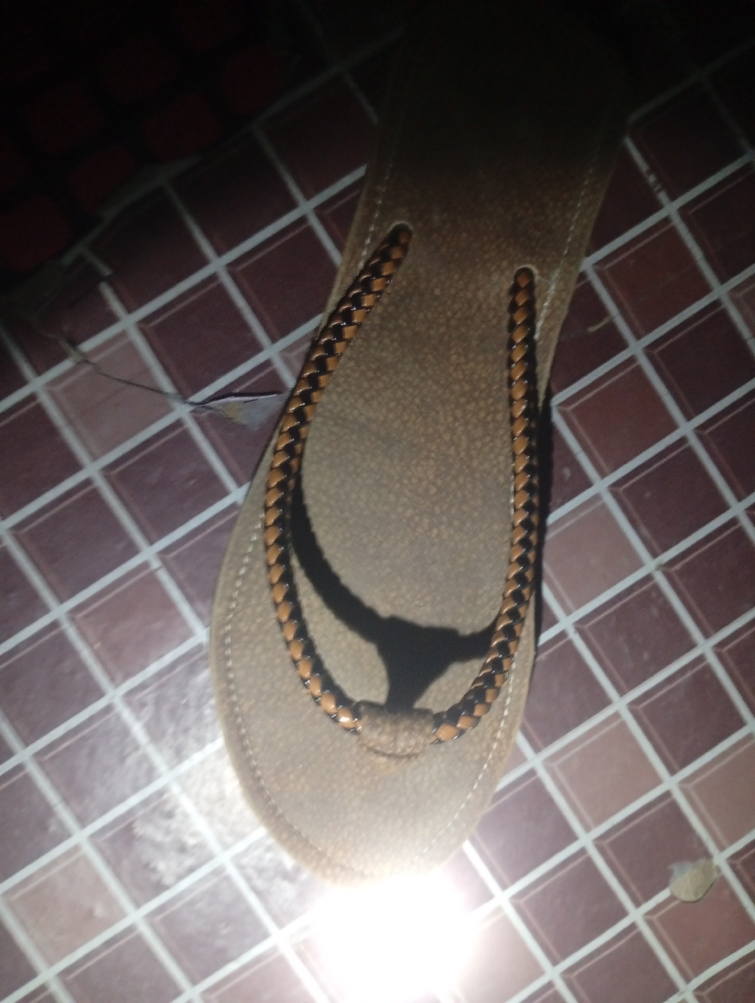 A close-up, overhead shot captures a single flip-flop lying on a tiled floor. The flip-flop is a neutral brown color with a woven strap made of alternating brown and black material. The strap forms a "Y" shape. The tiled floor has a pattern of square tiles in a reddish-brown hue with thin white grout lines. A shadow is cast on the flip-flop from an unseen light source. The lighting in the image is dim with a bright light source creating a glare on the bottom right side. The location is Unknown, Unknown.