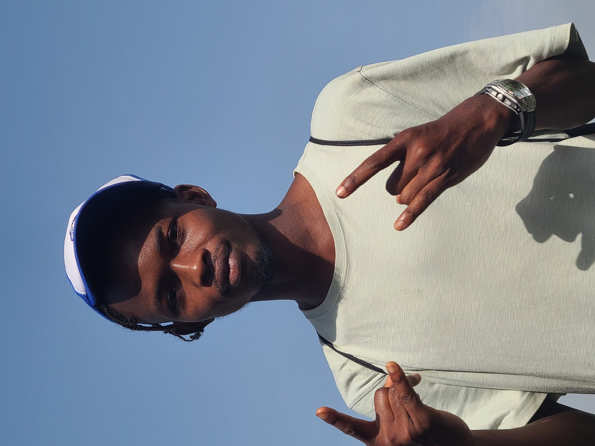 A person, viewed from a low angle against a clear blue sky, is making the "rock and roll" hand gesture with both hands. The individual is wearing a light green t-shirt and a blue and white baseball cap. The person has dark skin and appears to be wearing a watch or bracelet. The location in the city is unknown.