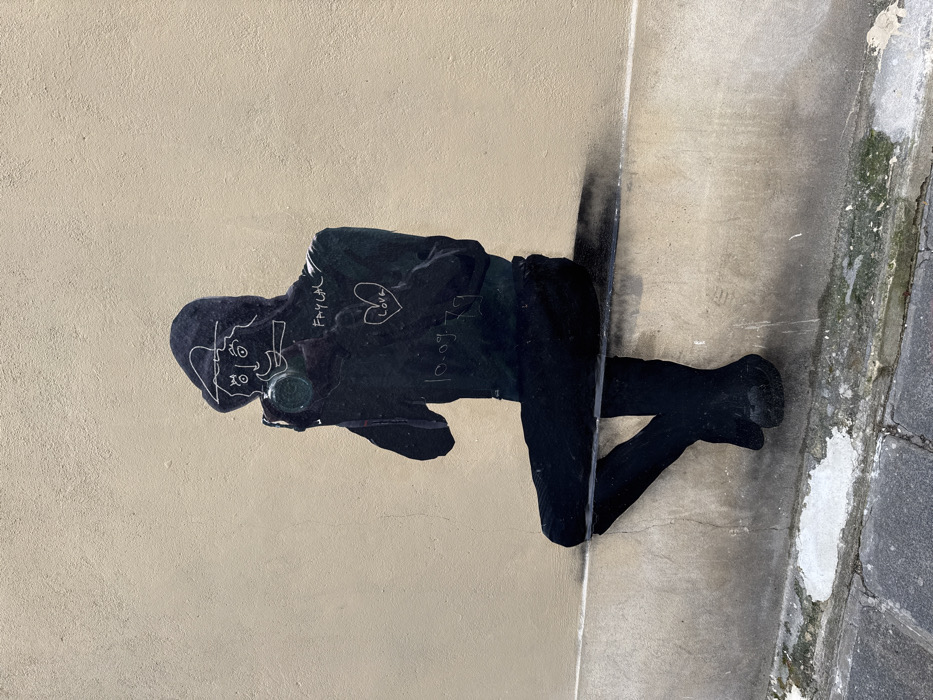 A piece of black street art, resembling a stencil or sticker, depicts a stylized, hooded figure sitting on a painted black ledge against a light beige wall in Paris, France. The figure is shown from the waist up, facing forward, holding a camera with a visible green lens reflection up to its face. White outlines on the dark hood delineate cartoonish facial features: two eyes, a nose, and a smiling mouth. Text applied in white over the figure's chest reads "FAYLAL," followed by a heart symbol enclosing "Love," and the date "10-09-79." The lower portion of the wall below the ledge exhibits discoloration, grime, and peeling paint. Below the wall, a paved surface, likely a sidewalk, is visible, composed of grey rectangular stones with some white marks.