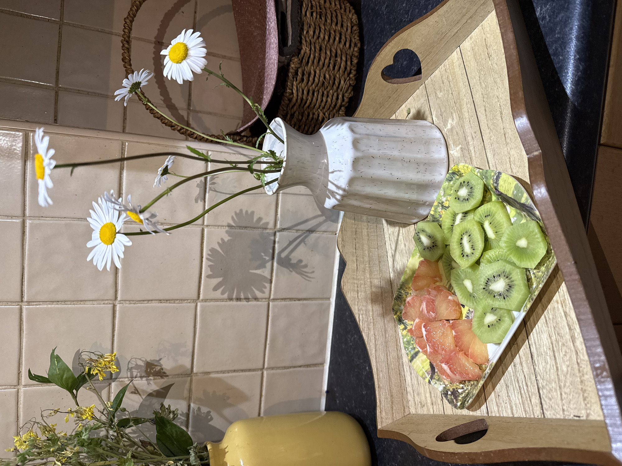 A still life arrangement on a countertop features a wooden tray holding a plate of sliced kiwi and grapefruit. A white vase containing daisies lies horizontally, with some flowers casting shadows on the tiled wall. A woven basket is in the background, and a yellow vase with greenery sits in the lower left corner.