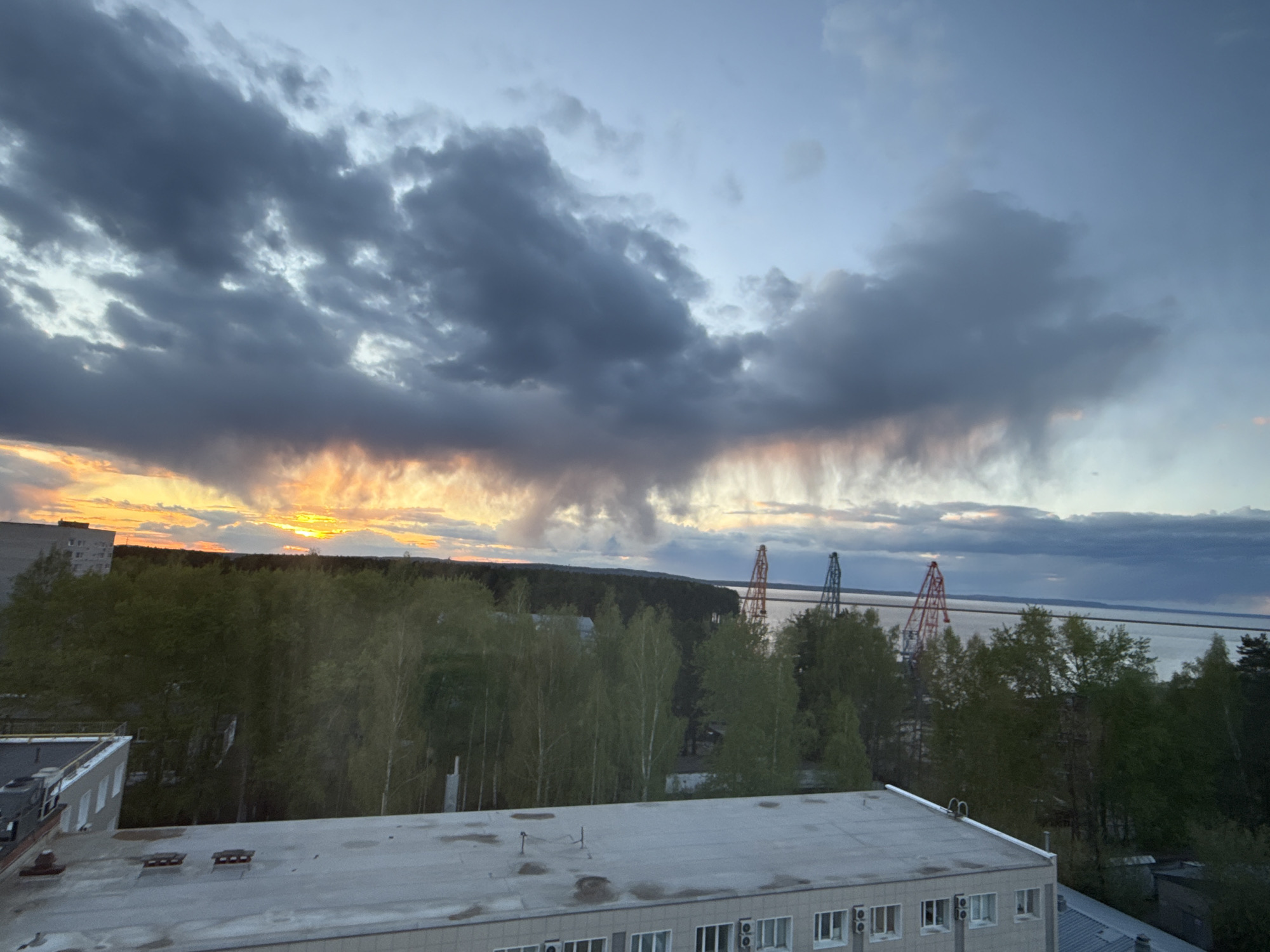 The image captures a cityscape at dusk or dawn. In the foreground, a flat, multi-story building with a light-colored roof dominates the lower portion of the frame. The building has visible air conditioning units installed near the windows.
Behind the building, a dense forest stretches across the midground. Interspersed within the trees are glimpses of other buildings, suggesting an urban-forest interface. Further back, near the horizon, there is a large body of water, possibly a river or lake.
Prominently visible amidst the trees are three cranes of different colors. The cranes are positioned on the water's edge, suggesting construction or industrial activity along the waterfront.
The sky is filled with dark, dramatic clouds, giving the scene a slightly ominous feel. There is a subtle orange glow on the left side of the image, likely due to the setting or rising sun.
Based on the provided location, it is plausible that the water is the Votkinsk Reservoir, and the cranes are part of an industrial facility or dockyard along its shores, close to the city of Chaikovsky.