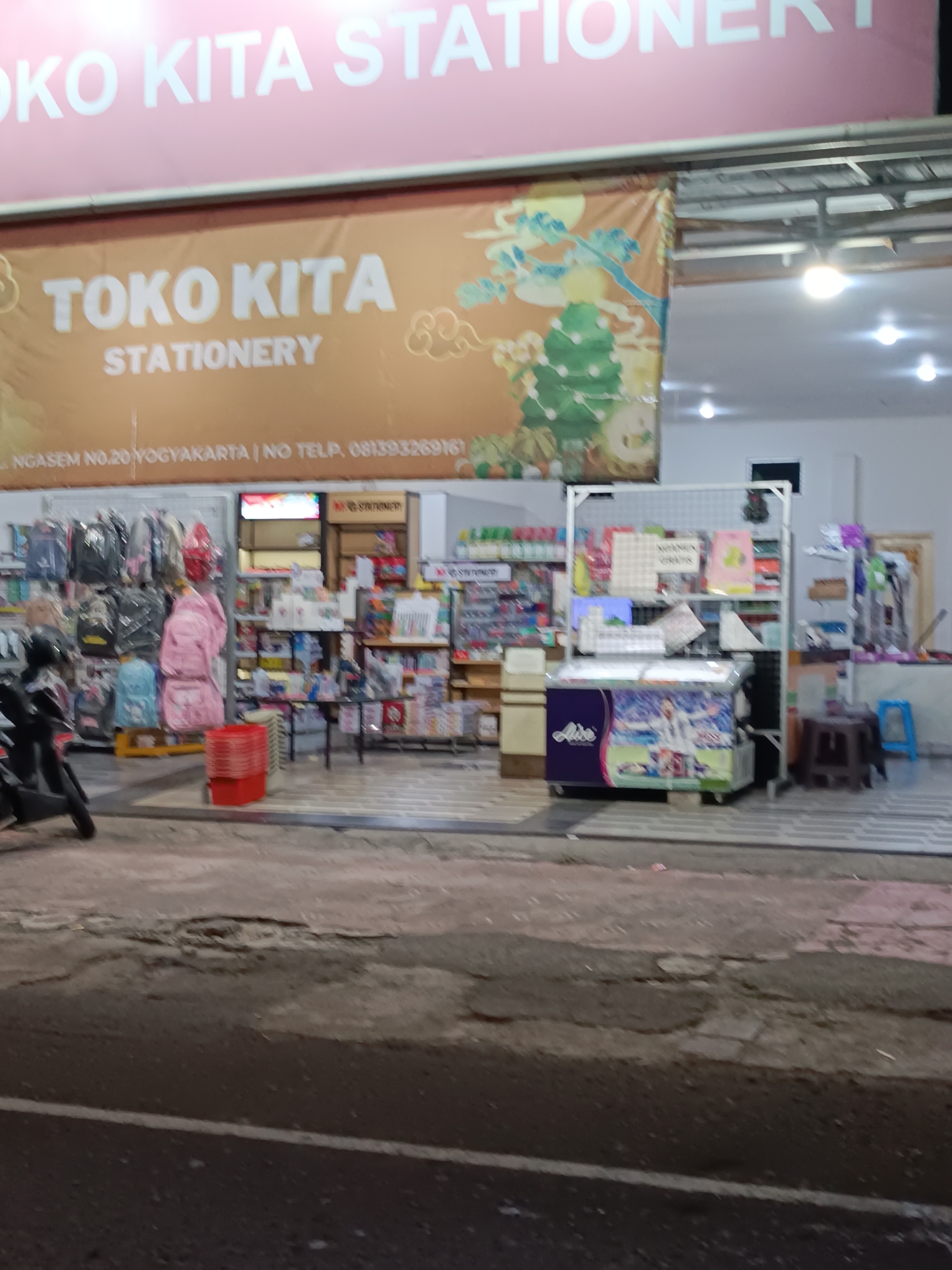 The image shows the storefront of "TOKO KITA STATIONERY" located at "NGASEM NO.20 YOGYAKARTA | NO TELP. 081393269161". The store has a prominent banner with the store name and a decorative design. Inside the store, shelves are filled with various stationery items, backpacks, and other merchandise. A refrigerator unit with colorful graphics is visible. The foreground shows a street with a motorcycle parked on the left. The overall scene depicts a commercial establishment in Yogyakarta, Indonesia.