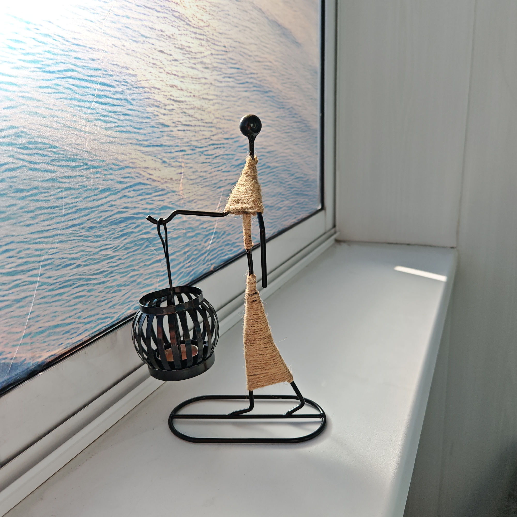 A metal and twine decorative object, possibly a candle holder, stands on a white windowsill. The object is shaped like a person carrying a lamp. The window is behind the object, displaying a blurred image of what appears to be a body of water, perhaps the Don River. The wall beside the windowsill is light grey. The location may be Rostov, Russia based on the water visible through the window and the presence of a Don River.