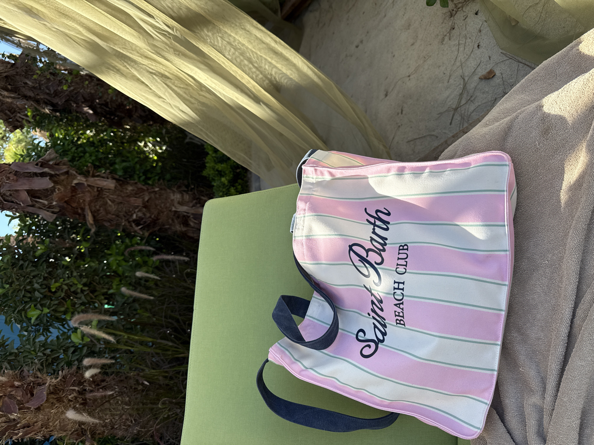 A pink and white striped canvas tote bag with "Saint Barth Beach Club" printed on it in black script and block letters rests on a folded taupe towel. The bag is positioned on a lime green cushioned lounge chair, which is situated outdoors. In the background, lush green foliage and palm trees are visible. A sheer, olive-green canopy or curtain drapes diagonally across the right side of the frame, casting shadows. The scene appears to be a sunny day, suggesting a vacation or resort setting. The overall impression is one of relaxation and summer leisure.