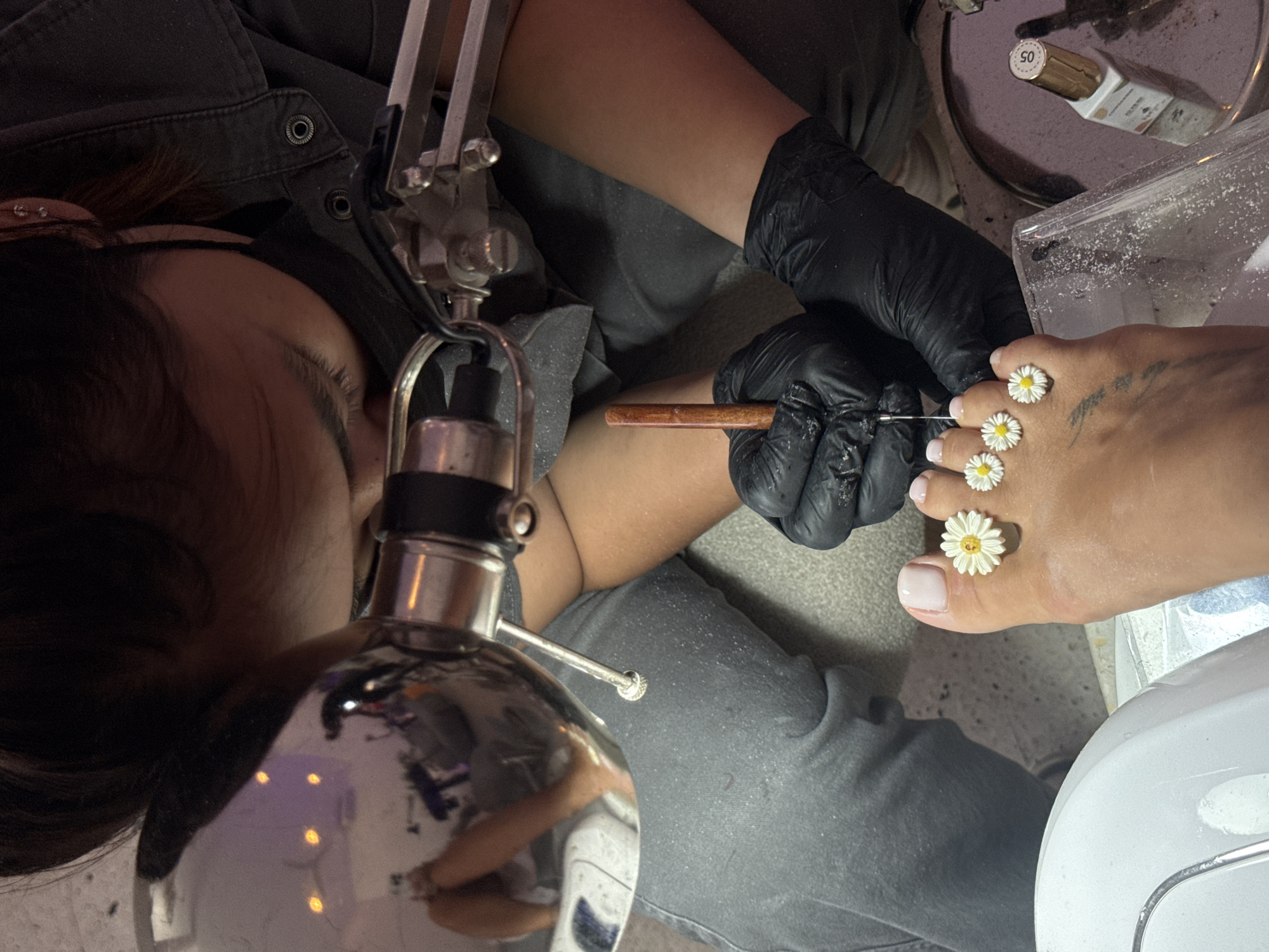This close-up, overhead shot captures a meticulous nail art application session, likely taking place in a salon in North Kuta, Indonesia. The image is dominated by the activity of a pedicurist, whose back of the head, shoulder, and gloved right arm are visible on the left and center. They wear a dark gray top and black protective gloves, suggesting a professional setting.
The focal point of the activity is the client's right foot, positioned on the right side of the frame. Four of the client's smaller toes are adorned with delicate, three-dimensional white daisy-shaped decorations, while the big toe showcases a perfectly applied light pink or nude nail polish. The pedicurist is actively engaged, holding a slender wooden tool in their gloved hand, indicating the precise work involved in attaching or refining the toe decorations.
Various objects related to nail care are scattered around the workspace. A prominent metallic task lamp with a flexible arm provides focused illumination from the left, its reflective surface showing blurred lights from the surroundings. In the upper right corner, a small bottle, possibly nail polish or a treatment product, is visible with the number "05" clearly printed on its cap. Beside it, a clear plastic container holds a white powdery substance, likely used for nail enhancements. A white UV/LED nail lamp is partially visible in the bottom right, typically used to cure nail products. The pedicurist's dark gray pant leg, seen below their arm, is covered in fine white dust, a common byproduct of nail filing or product application. The scene conveys a sense of focused attention and skillful execution during a beauty treatment.
