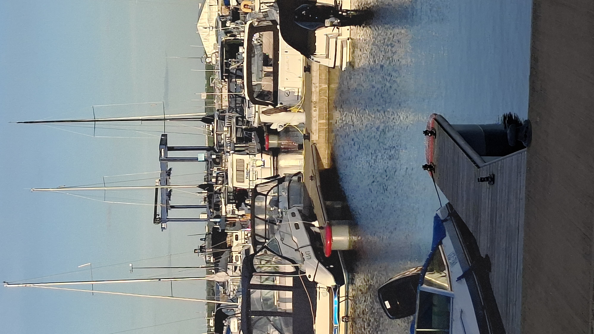 An outdoor scene depicts a marina with numerous boats docked in clear, blue water under a bright, pale blue sky. In the foreground, a wooden pier extends from the right side of the image, with a small boat tied to it. The boat is white with a blue stripe and has an outboard motor. The pier has several cleats and a dark bollard at the corner.
In the midground, several motorboats and yachts are moored, some with canopies and cabins. One boat in the center has a white cabin with tinted windows and is labeled "TIGETTY". Another boat to the left has a black and white hull and is labeled "VL477". Red buoys are visible attached to the docks.
In the background, several sailboats with tall masts are docked, their sails furled. A blue gantry crane stands prominently behind some of the boats. In the far background, a white building with a peaked roof is visible, along with other structures and greenery. The water reflects the sky and the boats. The overall impression is of a calm day at a well-maintained marina.