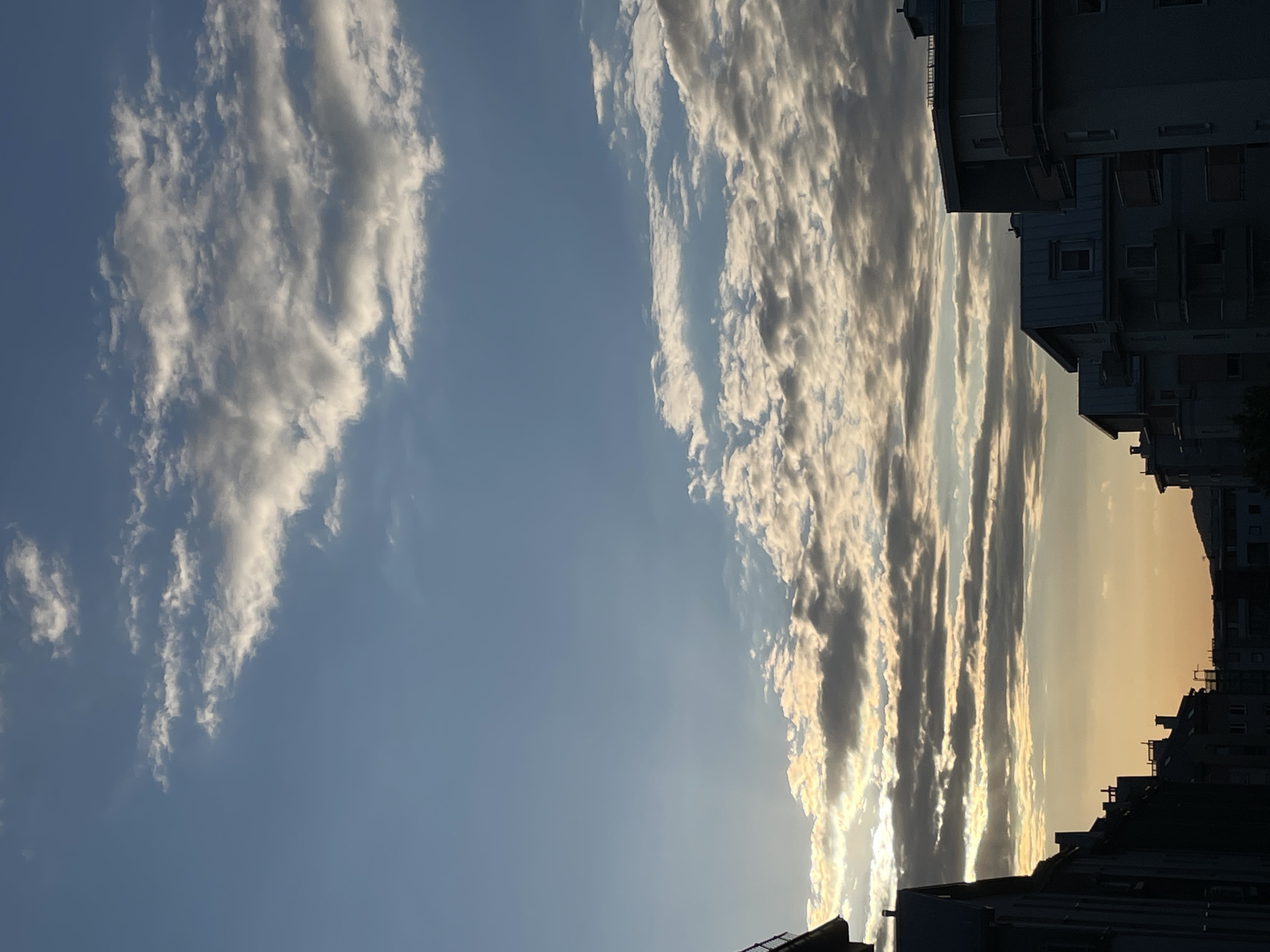 The image depicts a daytime scene with a sky dominated by clouds. On the left side of the frame, there are puffy white clouds scattered against a blue sky. Towards the right, the clouds appear more elongated and streaked, tinged with a golden hue, likely from the sun. In the lower right corner, the dark silhouettes of buildings suggest an urban setting. The image is oriented vertically. The city is unknown.