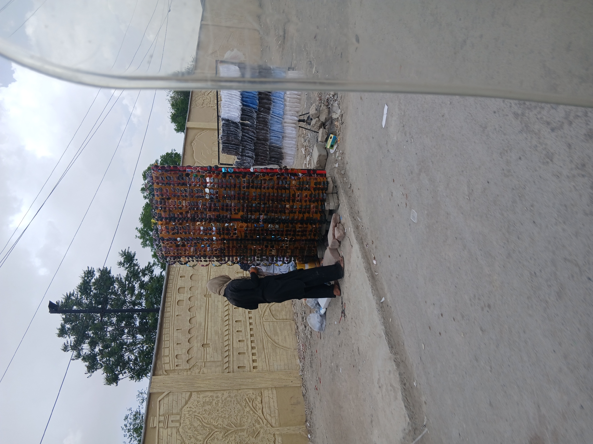 The image shows a street scene in Quetta City Tehsil, Pakistan. A man dressed in black stands next to a display rack filled with sunglasses, presumably a street vendor selling sunglasses. Behind the vendor is a wall with raised, decorative artwork. Adjacent to the sunglasses rack is another rack holding piles of folded clothing, likely also for sale. The street is paved and appears to be slightly dirty, with debris scattered along the edge. A utility pole is visible behind the wall. The shot seems to be taken from inside a vehicle, with the edge of the car window visible in the frame. The sky is overcast.