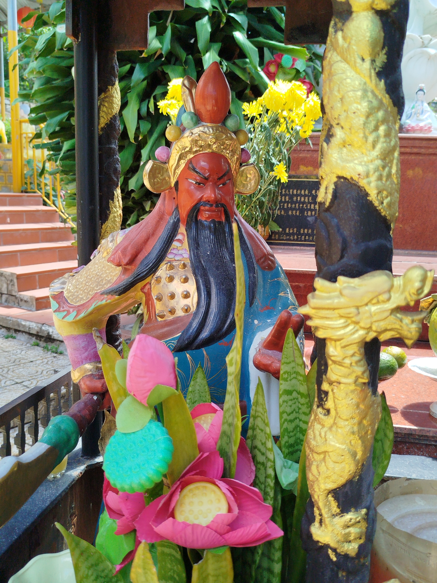 A close-up shot shows a weathered, painted statue of a bearded, red-faced deity with a long, black beard and headdress, possibly Guan Yu, placed at a temple in Da Lat, Vietnam. The statue is adorned with gold-colored ornamentation on its armor. Artificial lotus flowers and potted plants are placed around the statue. A black column featuring a gold-colored dragon design is to the right. Behind the statue, a building with steps, golden railings, yellow flowers, and text in Asian script is visible.