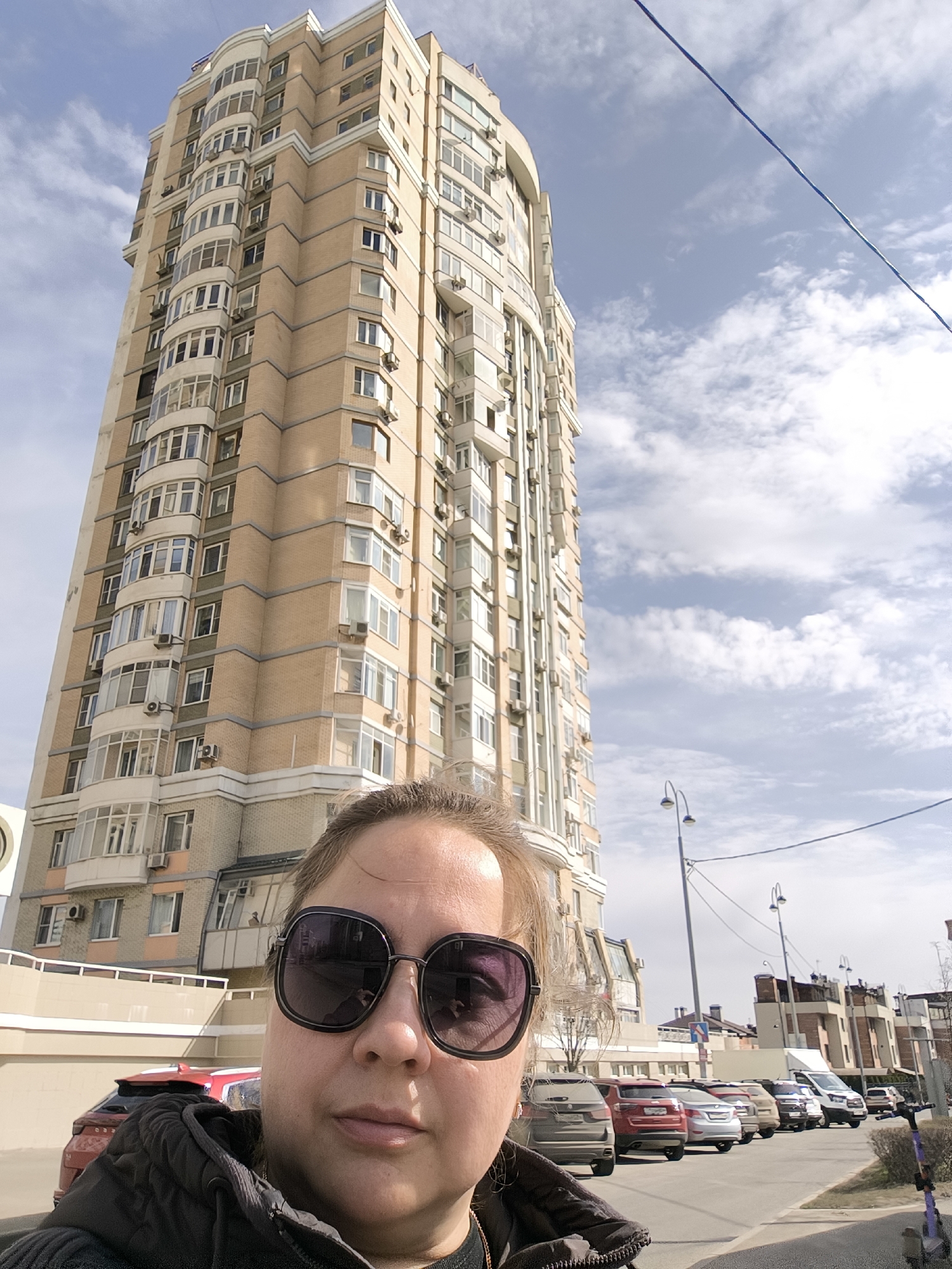A medium shot captures a woman taking a selfie in front of a tall building in Moscow, Russia. The woman wears oversized sunglasses and a dark jacket. Behind her rises a high-rise apartment building constructed of tan brick with white trim. Numerous windows and air conditioning units are visible on the building's facade. A row of parked cars lines the street adjacent to the building. Streetlights and buildings can be seen in the background across the street. The sky is partly cloudy.