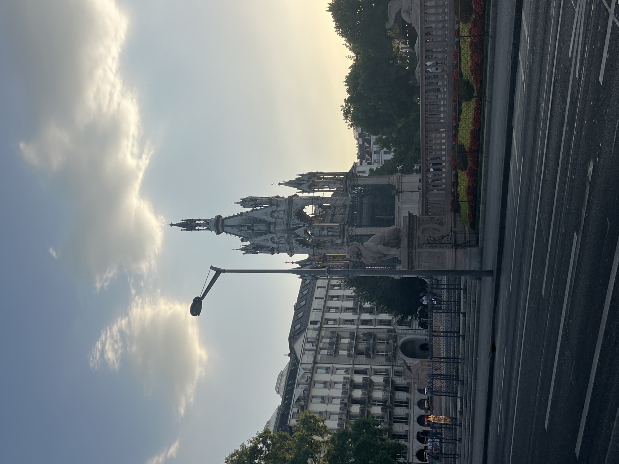 The image depicts a scene in Geneva, Switzerland, at the Brunswick Monument. In the foreground, a road with white lane markings stretches across the frame. To the right of the road, a lamp post stands tall. Behind the lamp post and to the right, the intricate Gothic architecture of the Brunswick Monument is visible, featuring spires and decorative elements, with a statue of a lion in front of it. To the left of the monument and across from it is a large, multi-story building with numerous windows and balconies, characteristic of European architecture. A metal fence separates the monument area from the street. In front of the fence, a row of meticulously arranged flower beds showcases vibrant red and yellow blooms. Behind the monument and building, mature trees provide a lush green backdrop. The sky above is a pale blue, with scattered clouds, and the setting sun casts a warm glow, suggesting it is late afternoon or early evening. Several people are visible in the background, walking and standing, appearing to be tourists or locals enjoying the area.