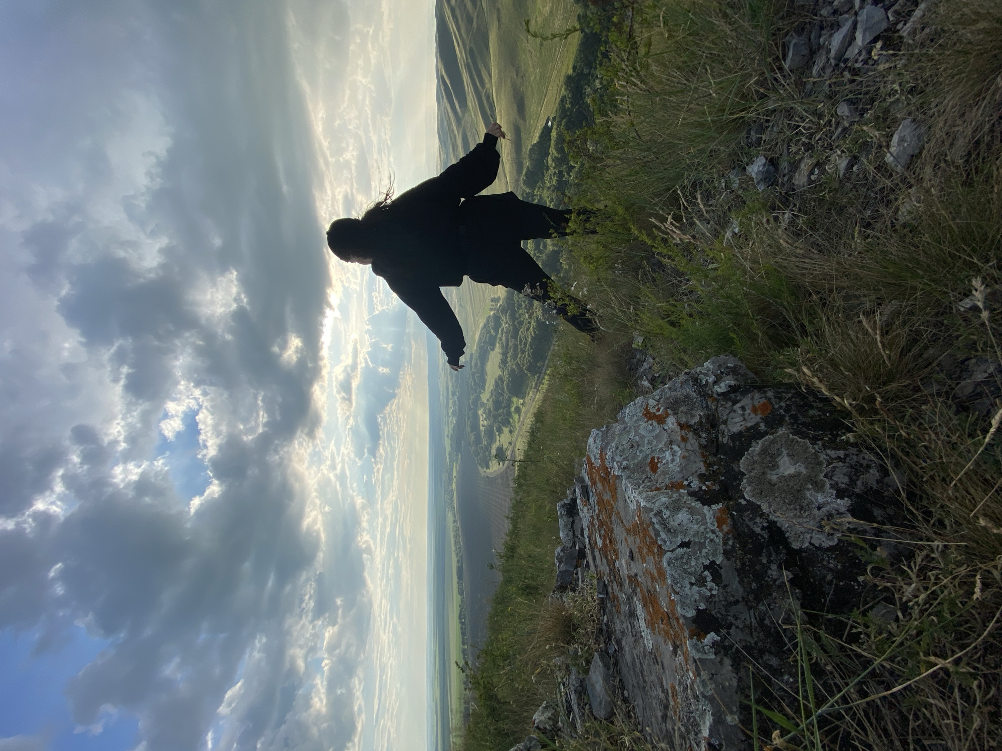 An overhead shot captures a young woman in silhouette, standing on a grassy precipice with her arms outstretched. She faces away from the camera, looking out over a vast valley bathed in the golden light of a setting sun. The sky above is a dramatic canvas of dark, swirling clouds, broken by shafts of light that pierce through, illuminating patches of the distant landscape. Below, the valley floor is a patchwork of green fields and winding roads, with rolling hills receding into the hazy horizon. In the foreground, a large, moss-covered rock adds a sense of ruggedness to the scene. The overall impression is one of awe and serenity, with the woman appearing to embrace the expansive natural beauty around her.